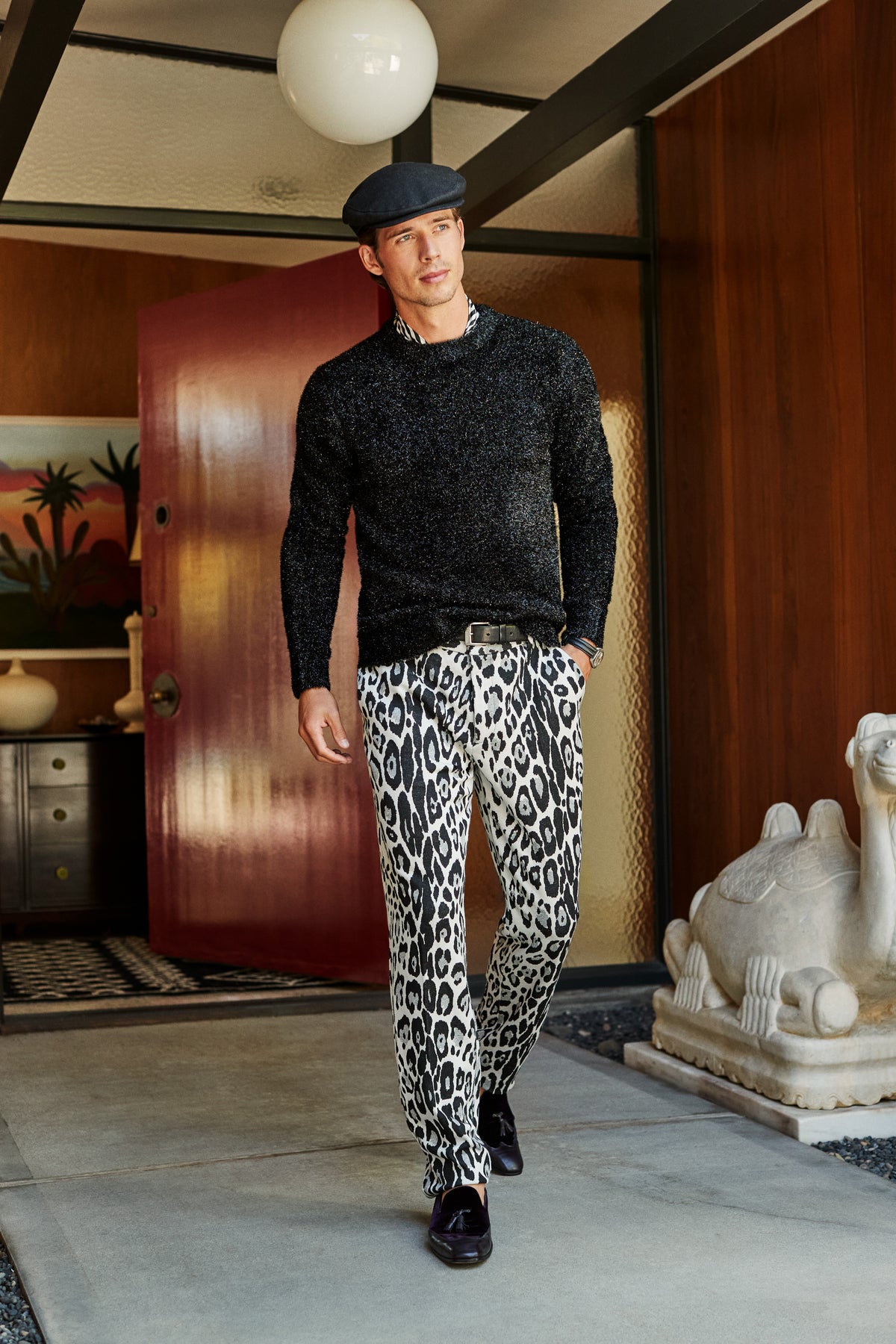 A man wearing a black sweater and leopard-print Clyde Slim Trouser walks out of a doorway.