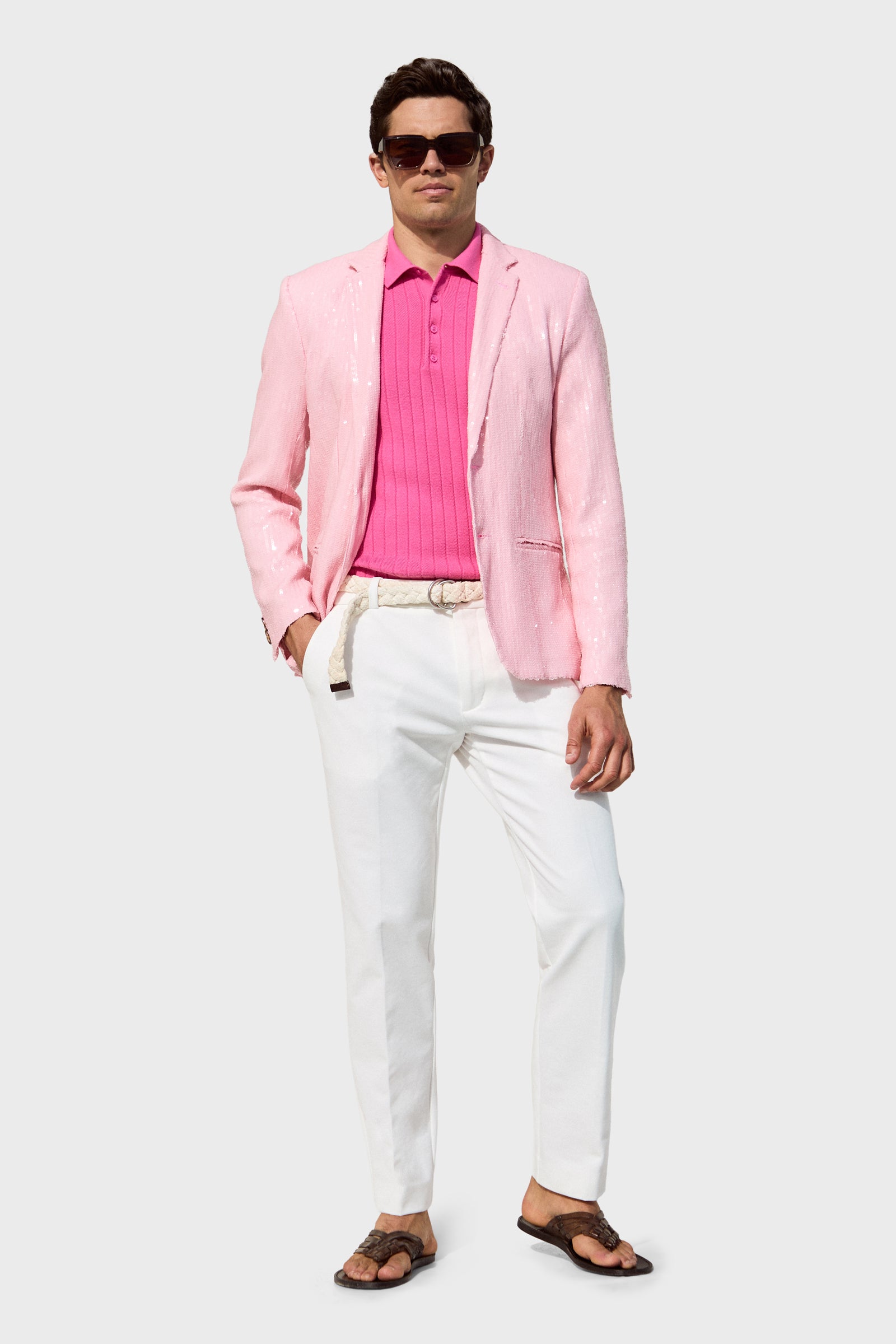 A man wears the Bowie Sequin Blazer with dark sunglasses, a bright pink polo, white pants, a beige belt, and brown sandals, standing with one hand in his pocket against a plain light gray background.