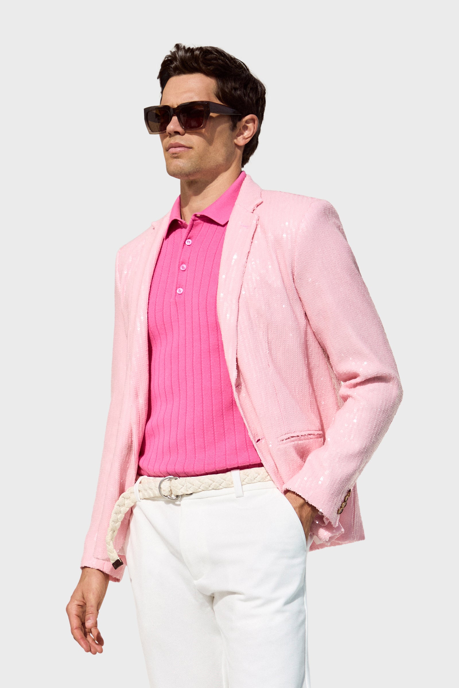 A man in dark sunglasses, a pink Bowie Sequin Blazer, a bright pink polo shirt, white trousers, and a white braided belt stands against a plain light gray background.