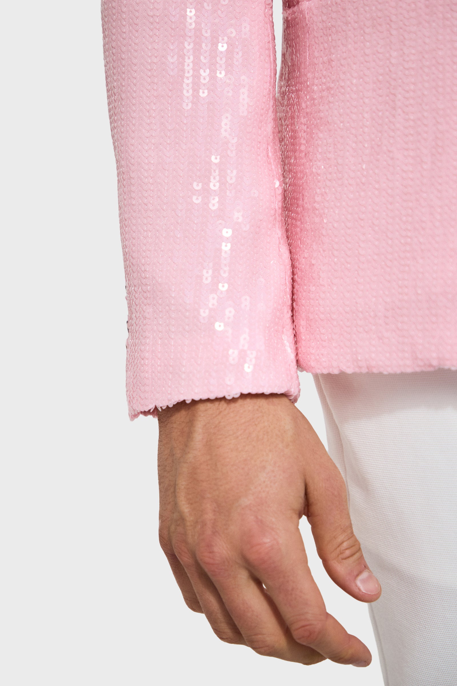 A close-up of a person's hand and forearm wearing the Bowie Sequin Blazer in pink, paired with white pants against a light gray background.
