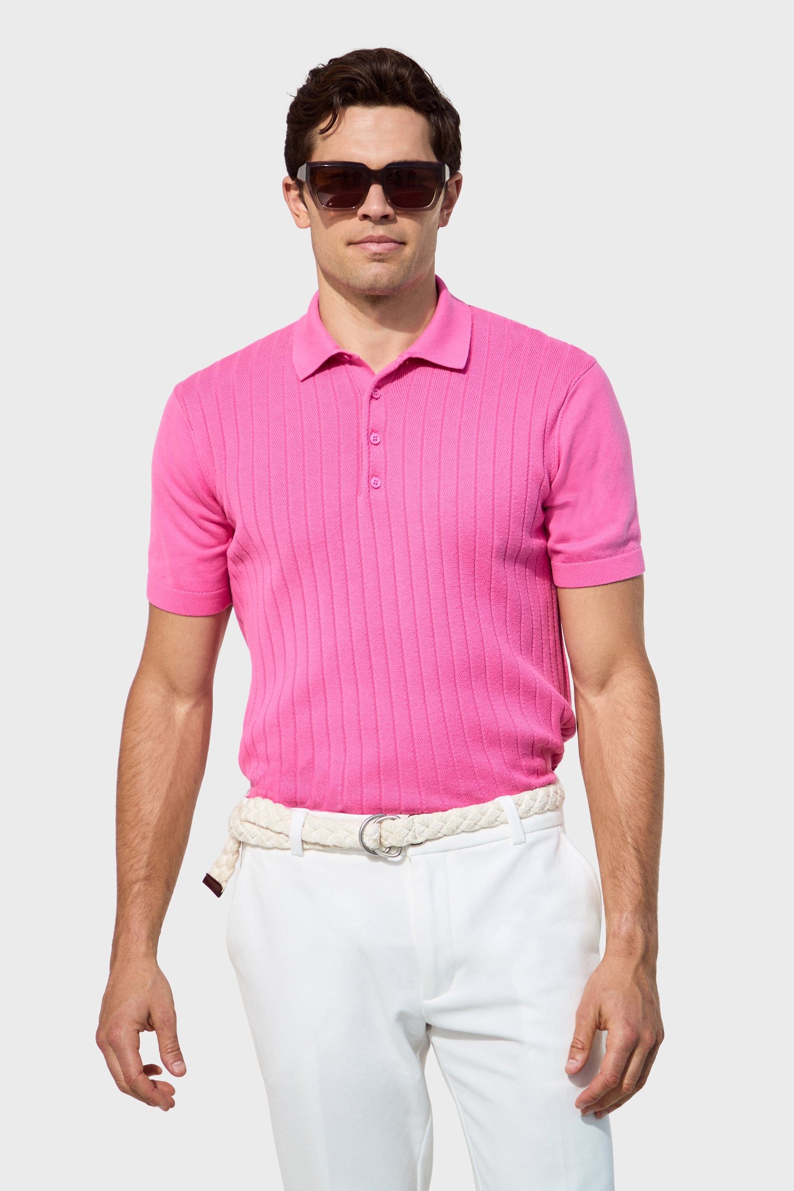 A man in dark sunglasses and the Alvarez Short-Sleeve Polo in bright pink, paired with white pants and a braided belt, stands against a plain light background.