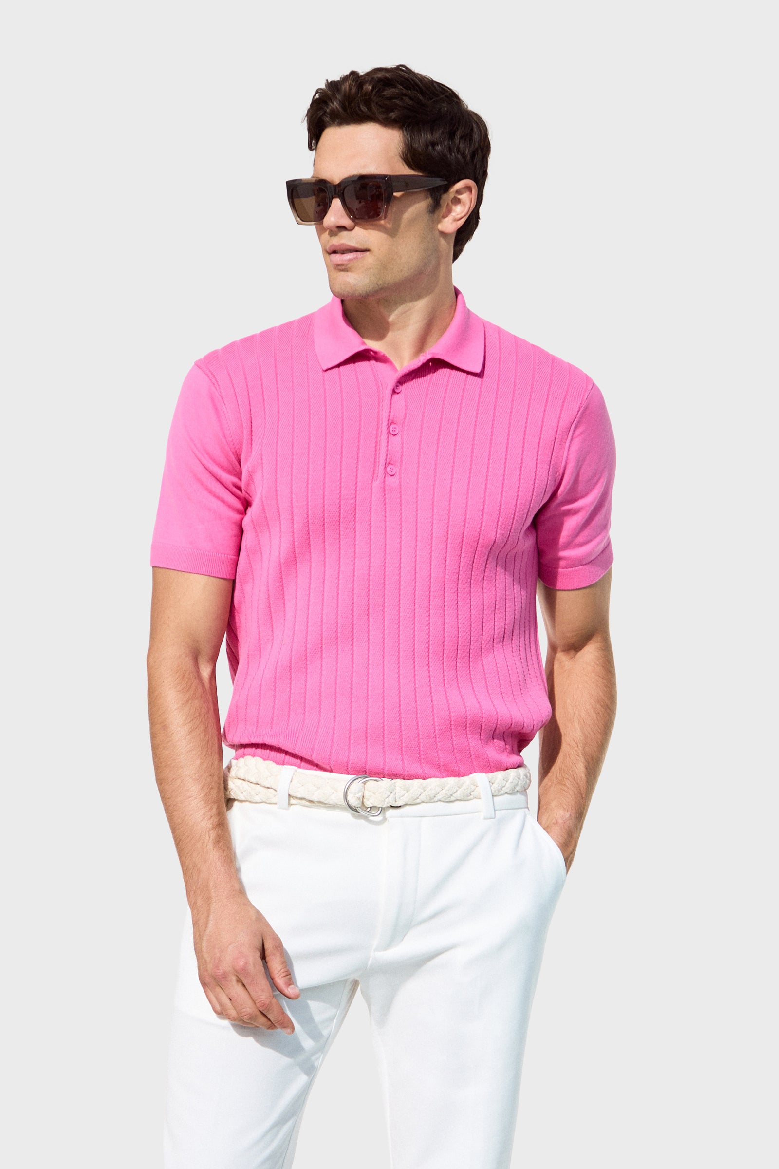A man in dark sunglasses, the bright pink Alvarez Short-Sleeve Polo, and white pants with a braided white belt stands against a light gray background.