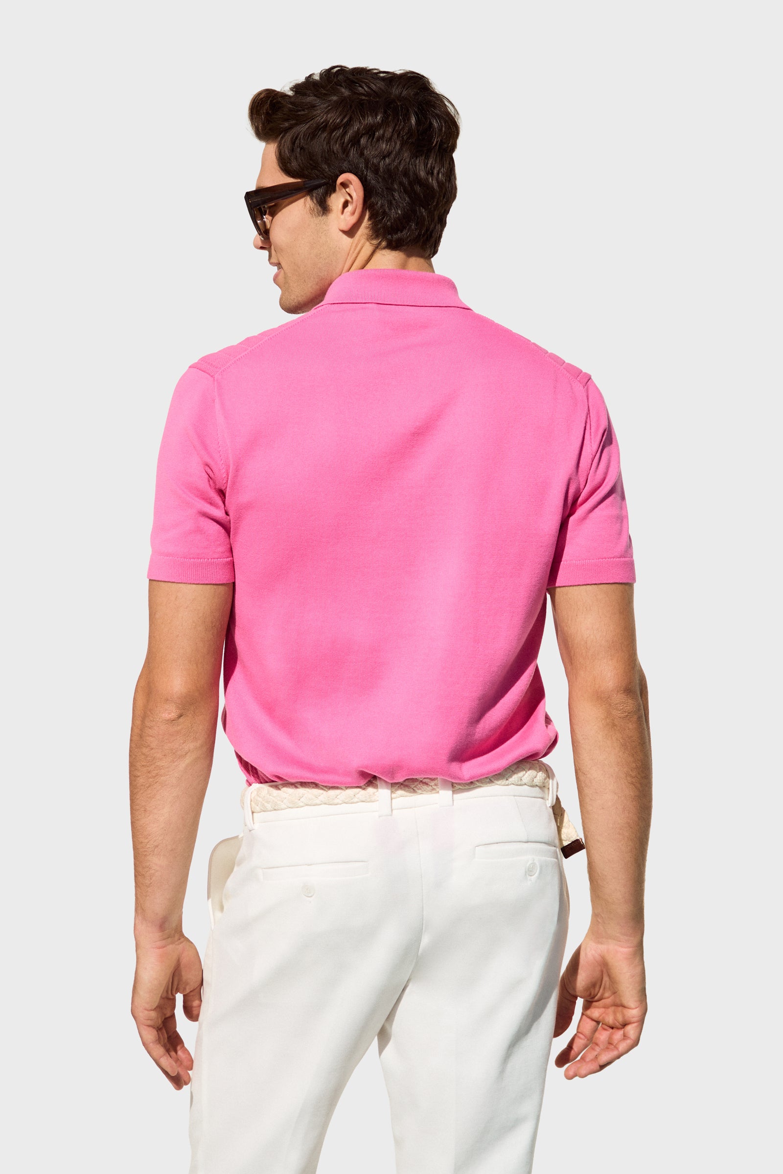 A man with short brown hair and sunglasses stands facing away, hands at his sides, wearing the bright pink Alvarez Short-Sleeve Polo and white pants. The background is plain light gray.