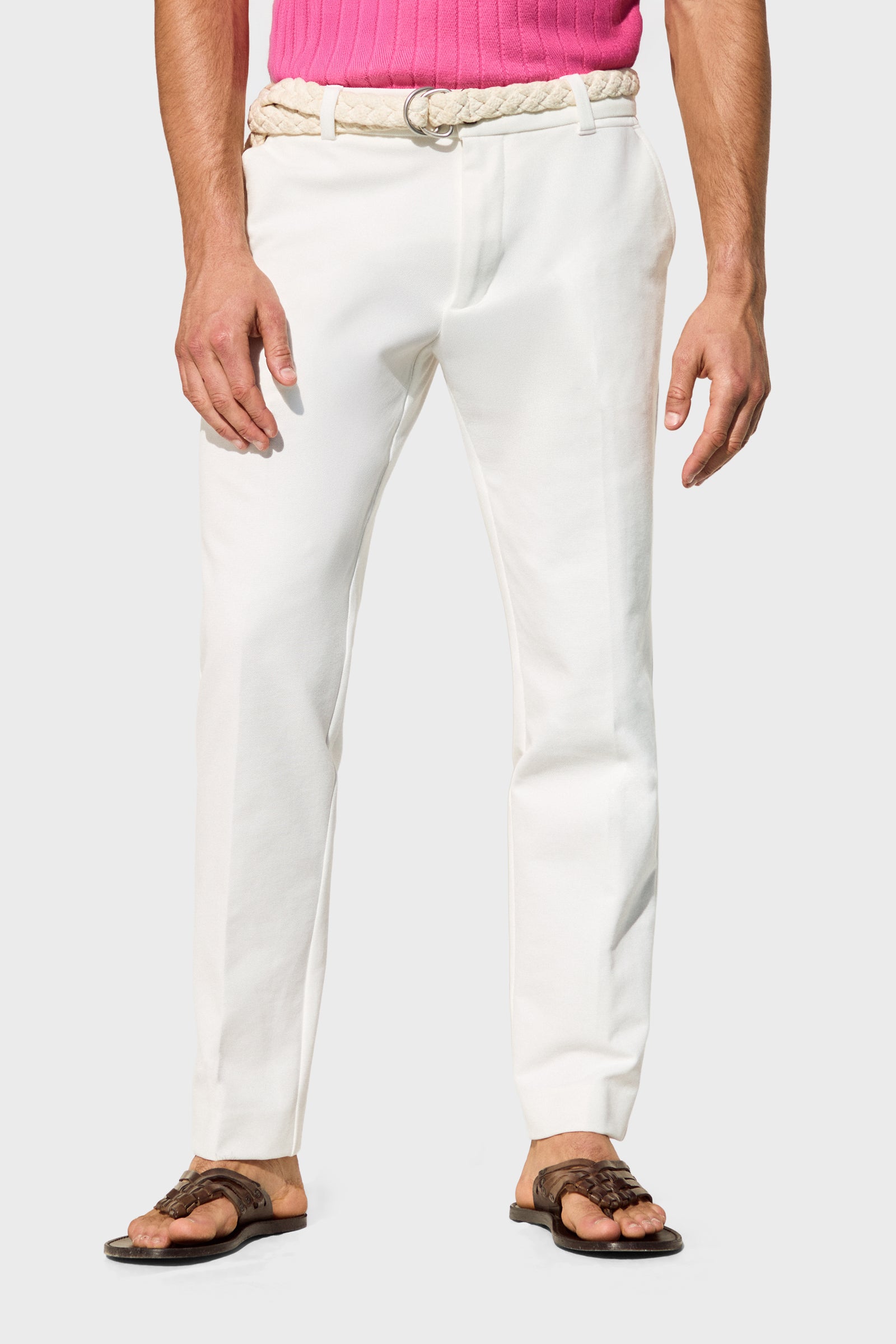 Someone wearing the Clyde Slim Trouser - Stretch Piqué with a braided belt, a partially visible pink shirt, and brown sandals stands against a plain light background.