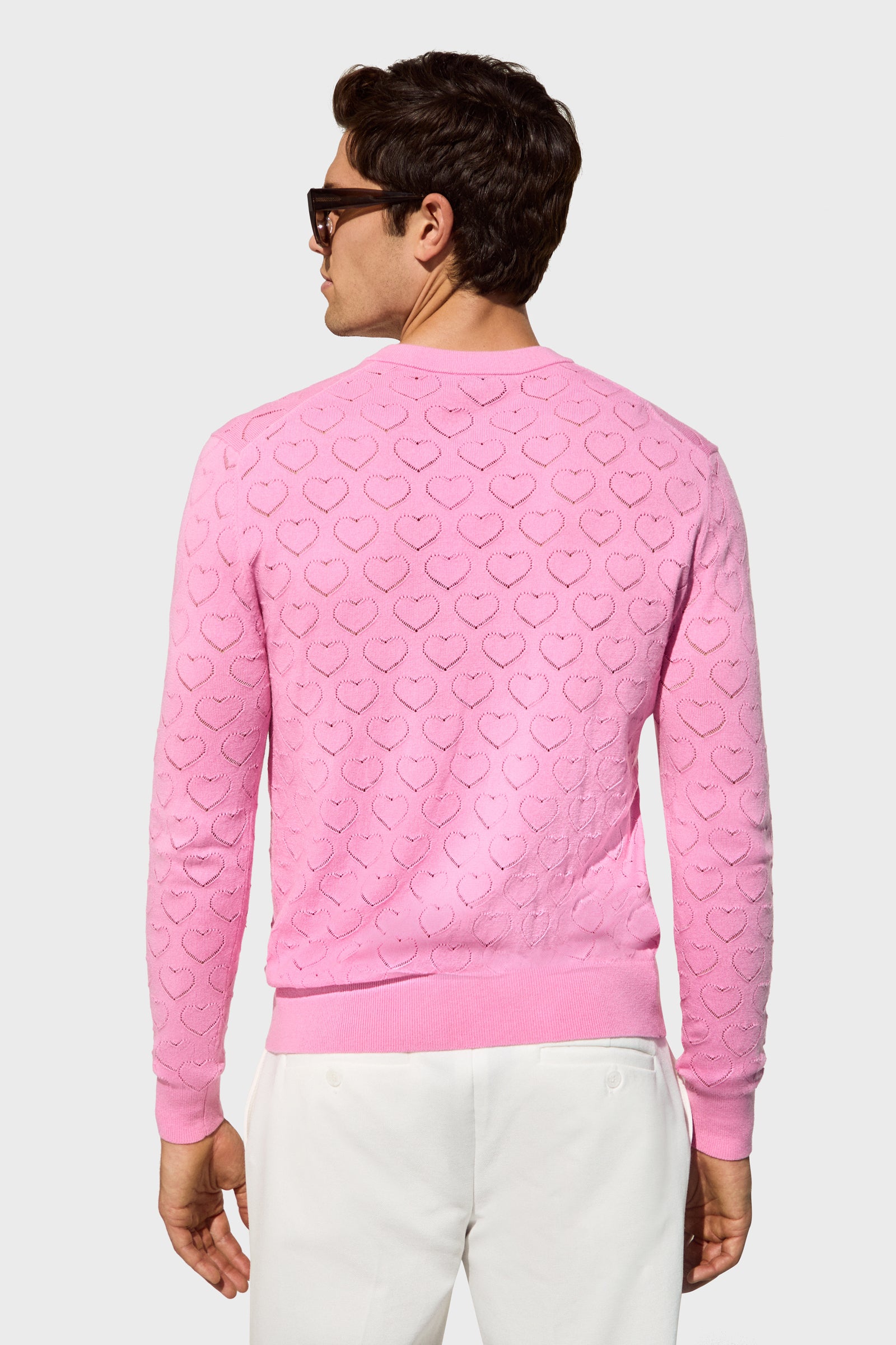 A man is seen from behind wearing dark sunglasses and the Eros Heart Pointelle light pink sweater, featuring a textured heart pattern, paired with white pants against a plain light background.