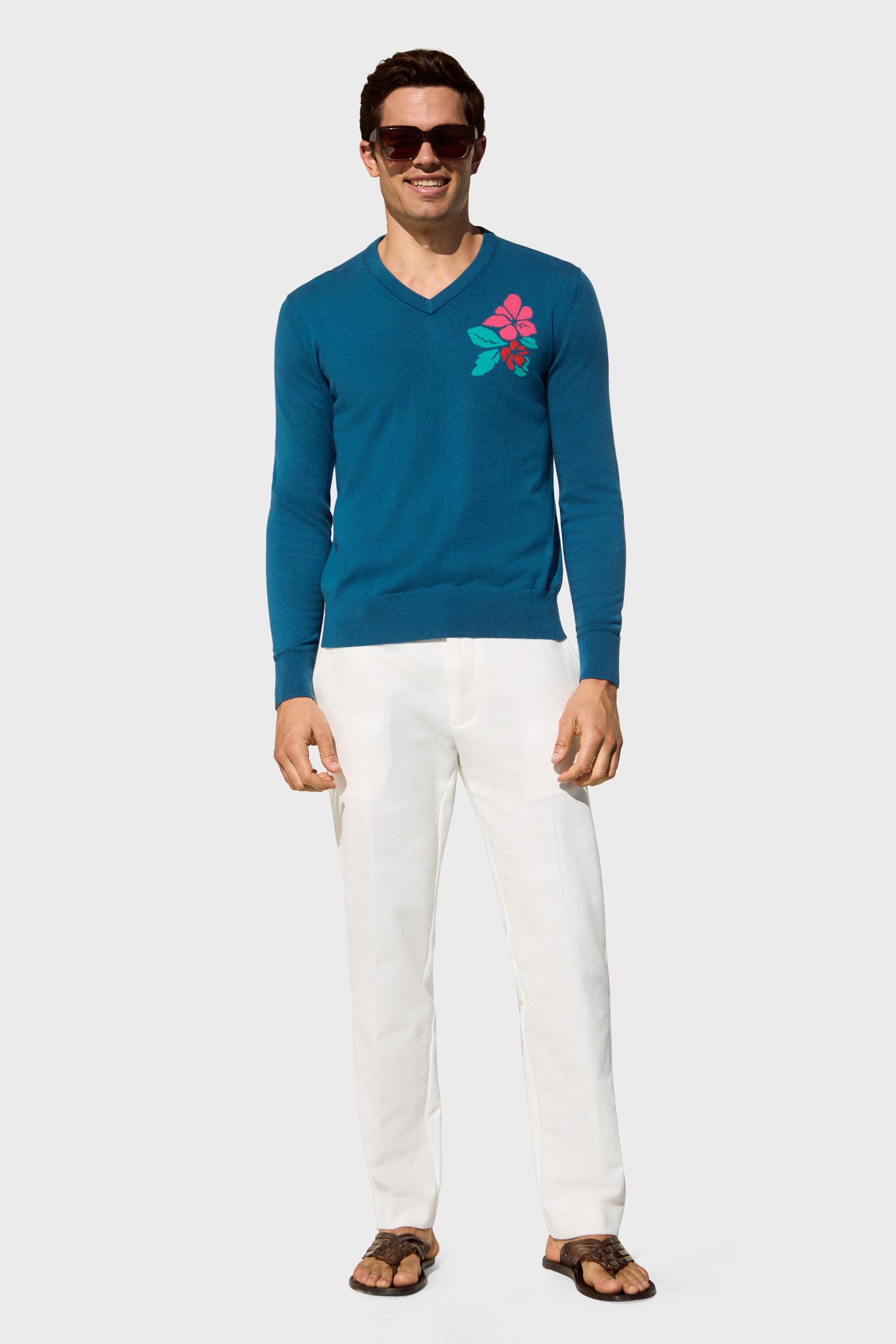 A man in sunglasses, white pants, and brown sandals smiles against a plain light gray background while wearing the Copacabana V-Neck Sweater featuring a pink and green floral design on the chest.