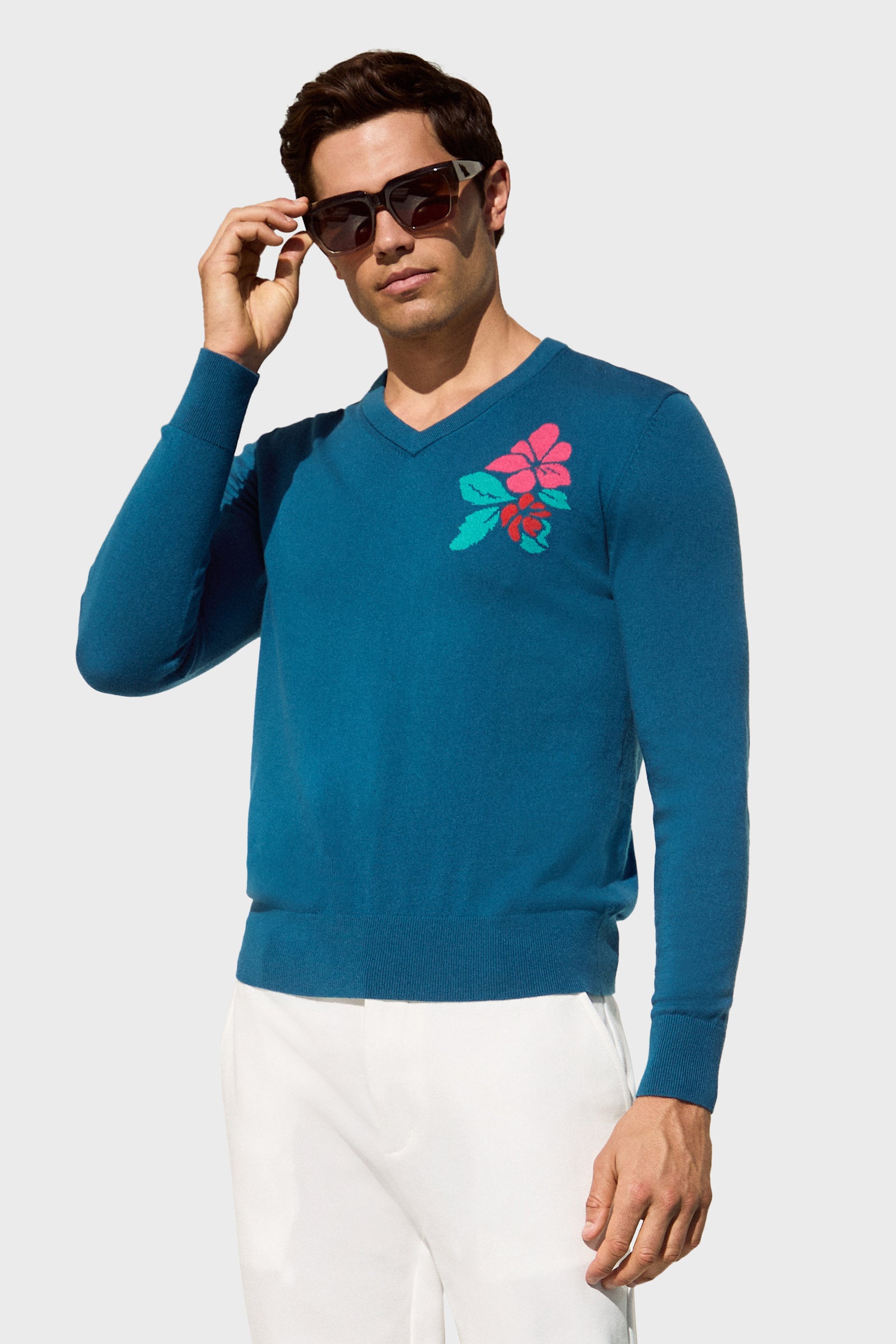 A man in dark sunglasses and white pants wears the Copacabana V-Neck Sweater, featuring a blue color with a pink flower on the chest, while standing against a light gray background.
