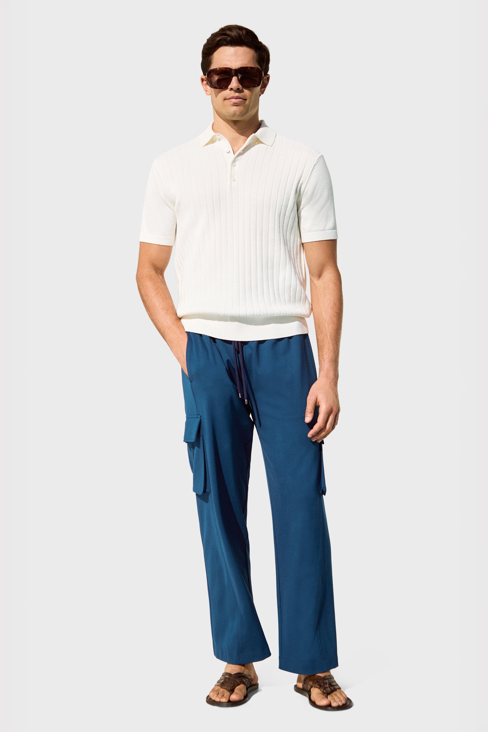 A man wearing sunglasses, the Alvarez Short-Sleeve Polo in white, blue cargo drawstring pants, and brown sandals stands against a plain light gray background.
