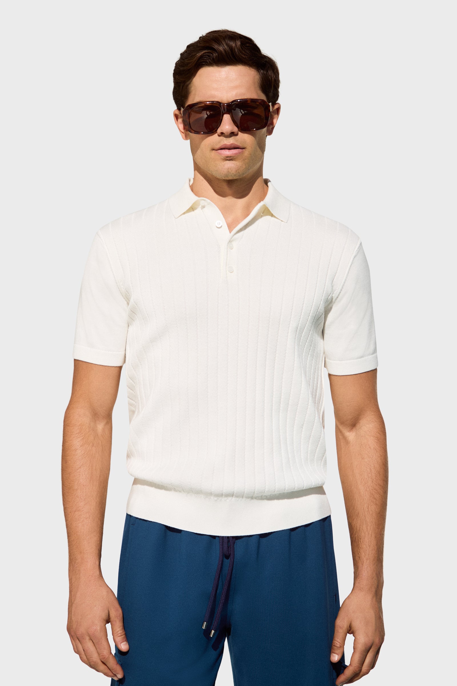 A man wearing dark sunglasses, an Alvarez Short-Sleeve Polo in white, and blue drawstring pants stands against a plain light gray background.