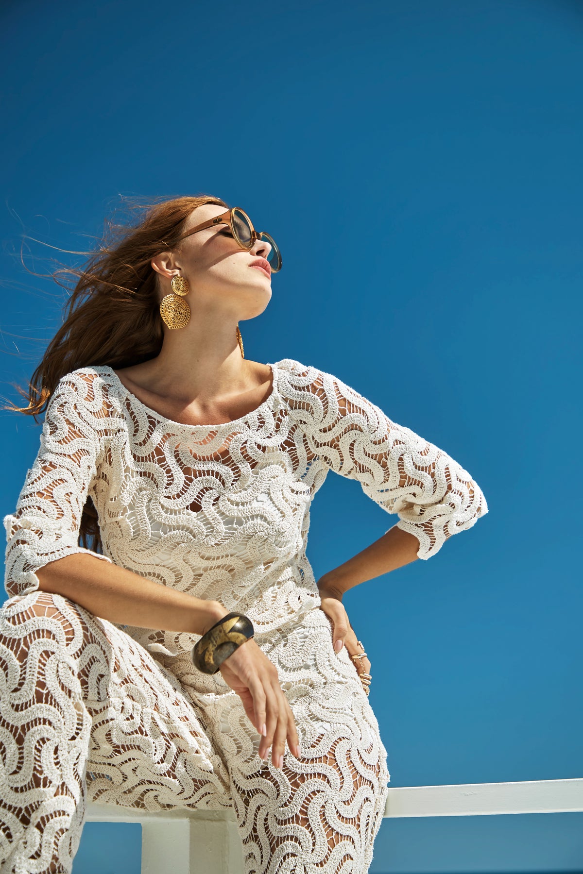A woman in sunglasses and the Triennale Top poses outdoors against a clear blue sky. She accessorizes with large earrings, a chunky bracelet, and her hair flows freely in the wind.