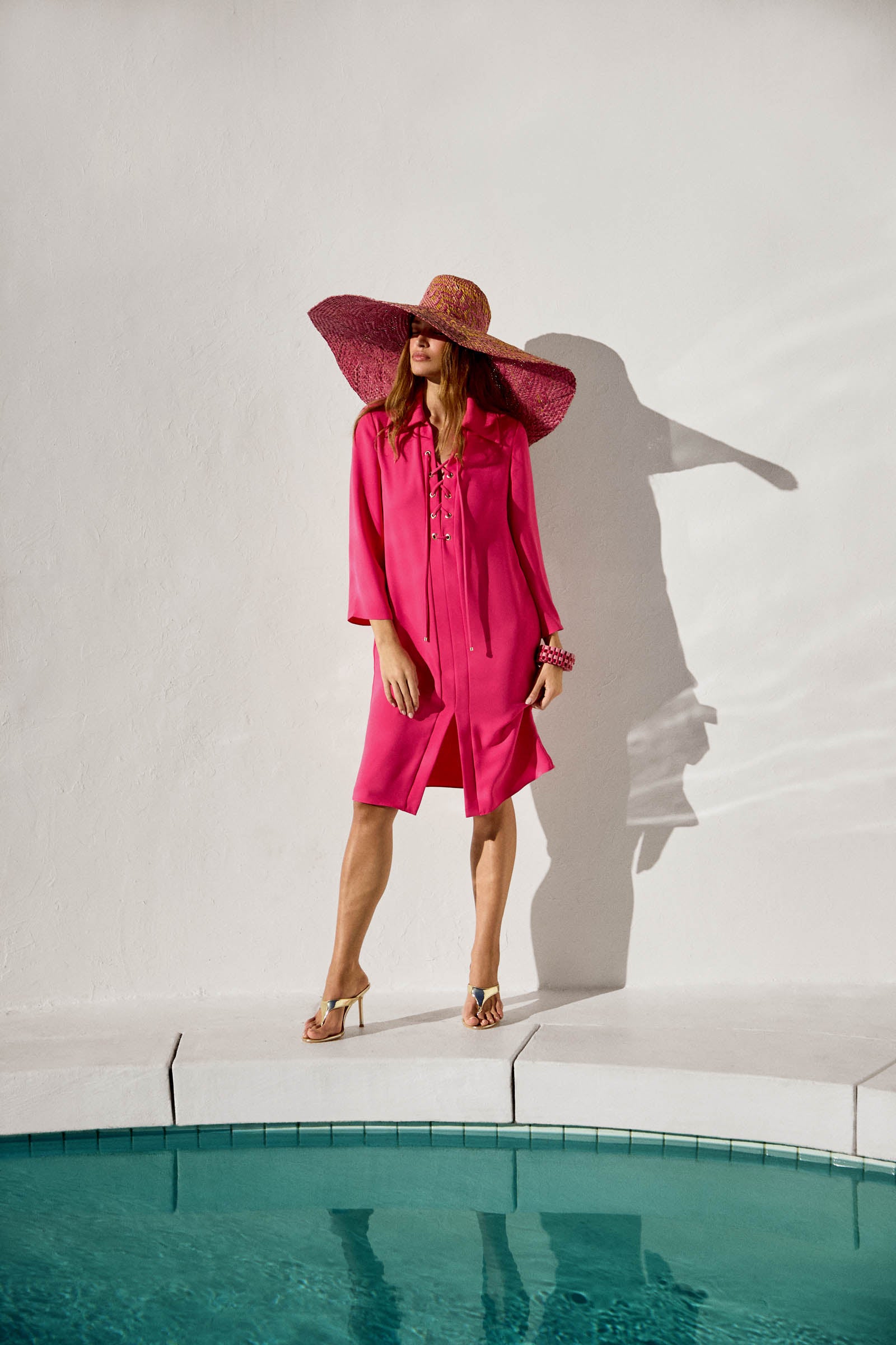 A woman in the vibrant pink Janeiro Lace-Up Dress and wide-brimmed hat stands by a pool, her silhouette casting a shadow on a white wall, accessorized with heeled sandals and bracelets for a chic, summery look.
