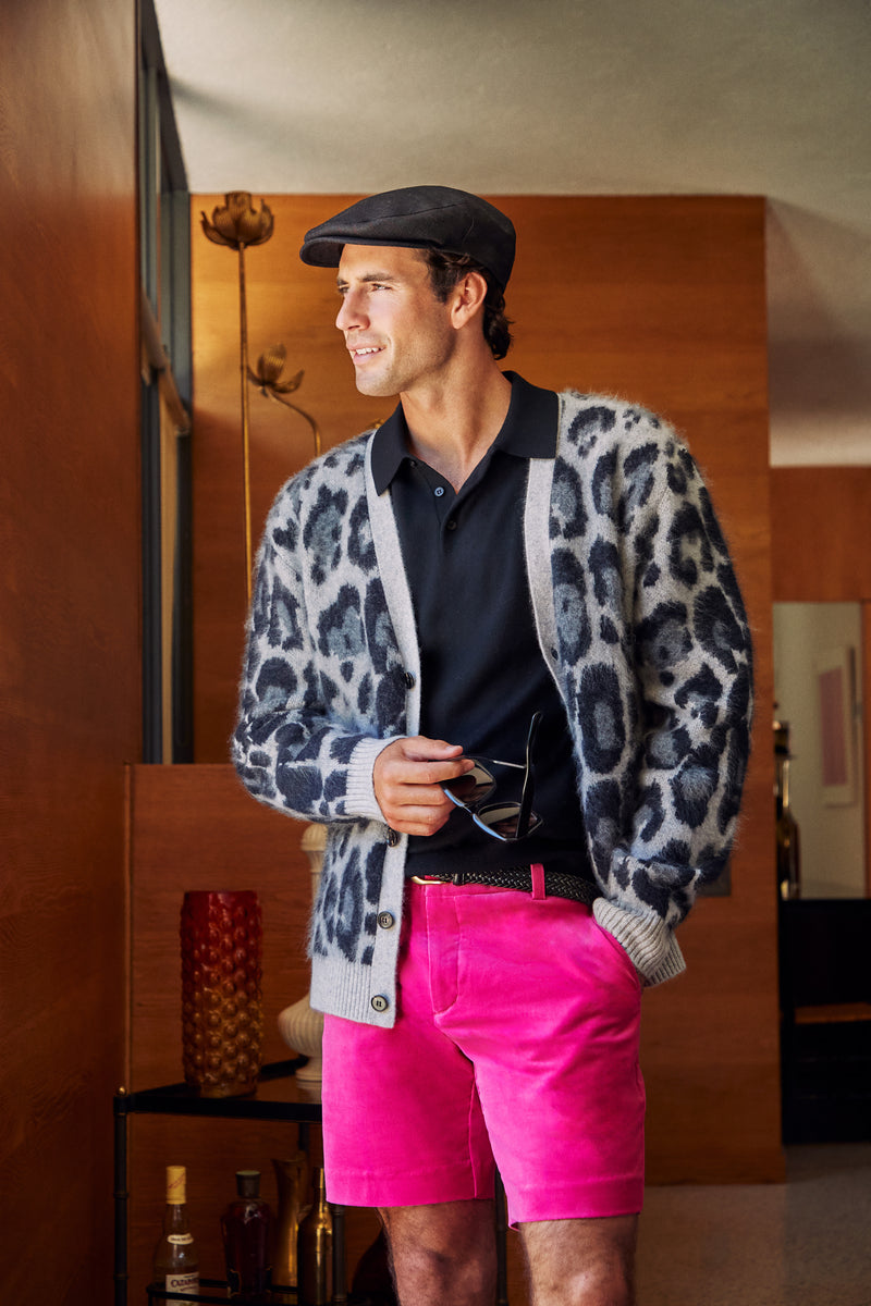 A man wears a LEO FUZZY CARDIGAN in grey leopard print over a black polo shirt and bright pink shorts, topped with a dark flat cap.