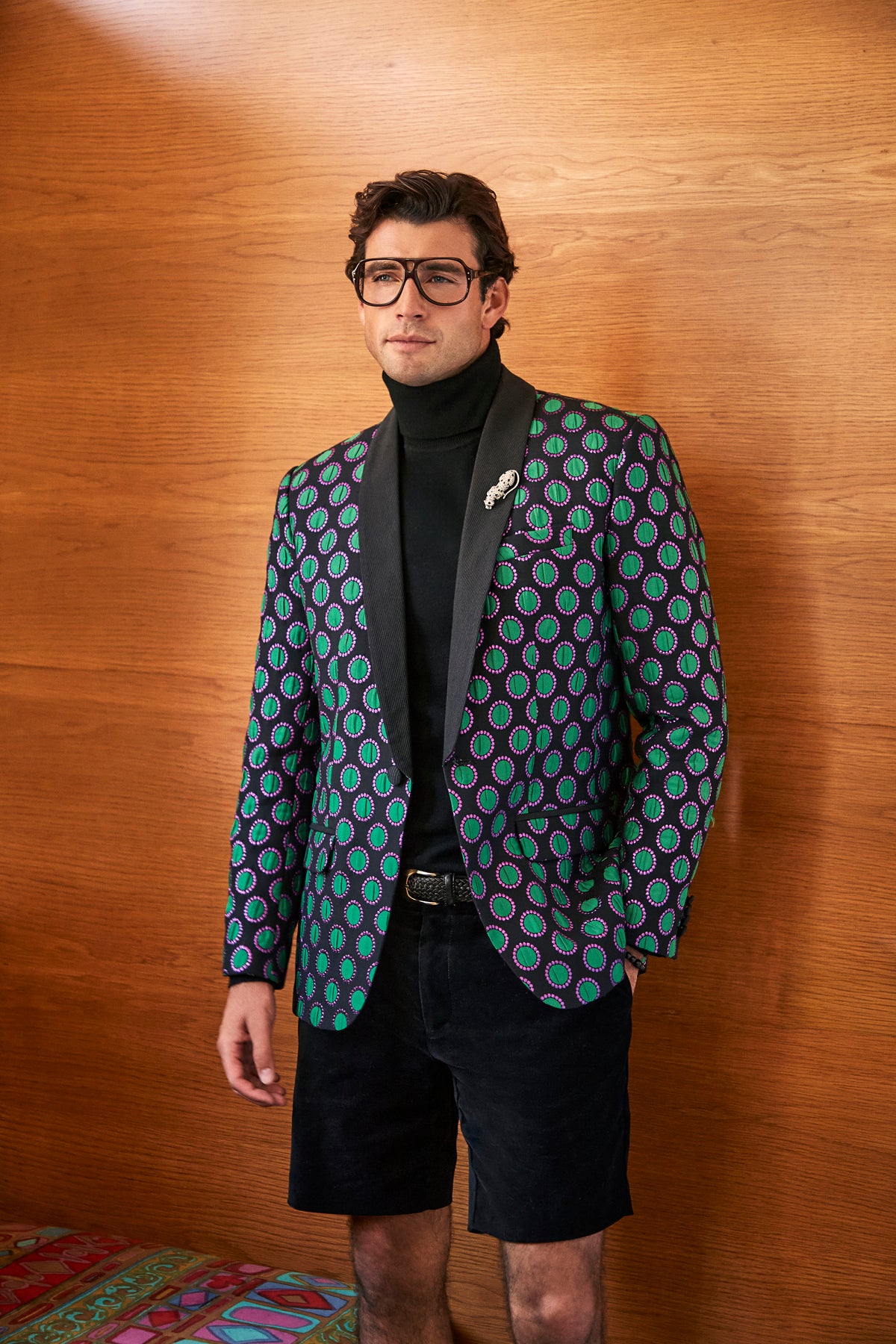 A man wears a black turtleneck and an Alfred Blazer in a green and purple geometric print, paired with black shorts.