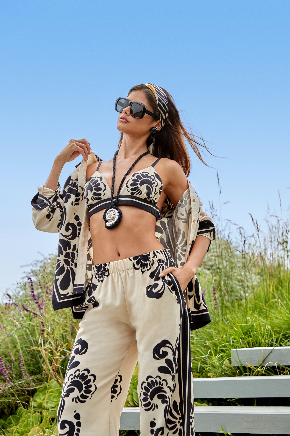 Wearing the Blanca Top, a woman in sunglasses and a patterned cream and black outfit poses outdoors by greenery and steps under a clear blue sky.