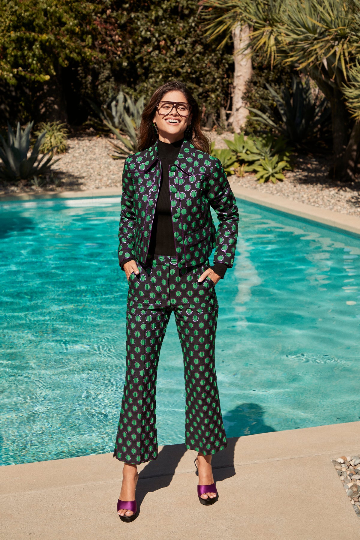 A smiling woman wearing a black turtleneck and an Allex Jacket with matching cropped trousers in a green circular pattern, standing by a swimming pool.