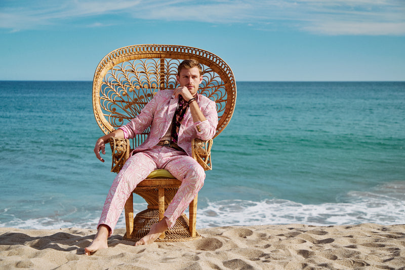 A man wearing the Thurston Blazer sits barefoot on a wicker chair on a sandy beach, with the blue ocean and sky in the background.