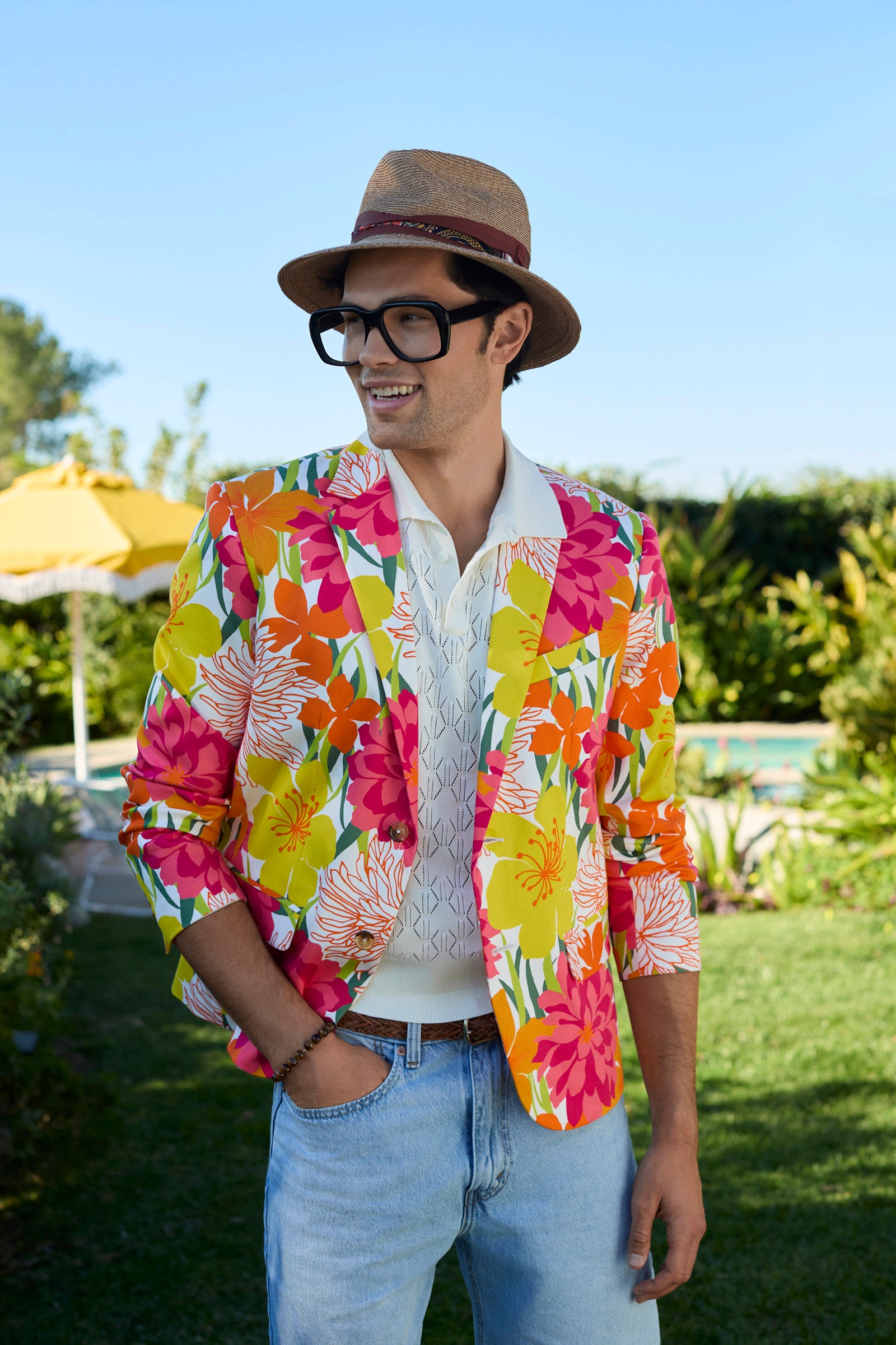 A man outdoors smiles in a sunny garden, wearing the Thurston Blazer - Como Herringbone over a light shirt and jeans, paired with a straw hat and large glasses. Green grass and yellow umbrellas are in the background.