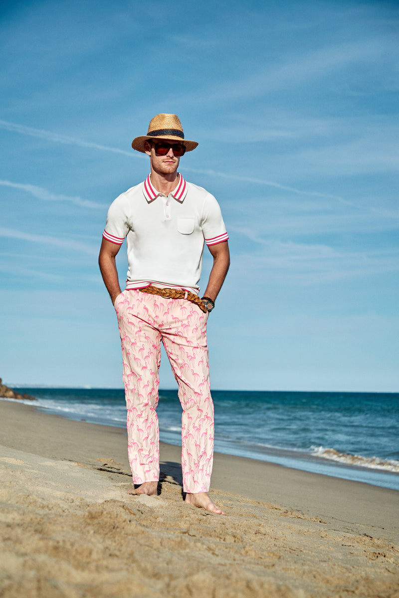A man stands barefoot on a sandy beach, wearing the Clyde Slim Trouser, a straw hat, sunglasses, and a white polo shirt with red trim, with the ocean and blue sky in the background.