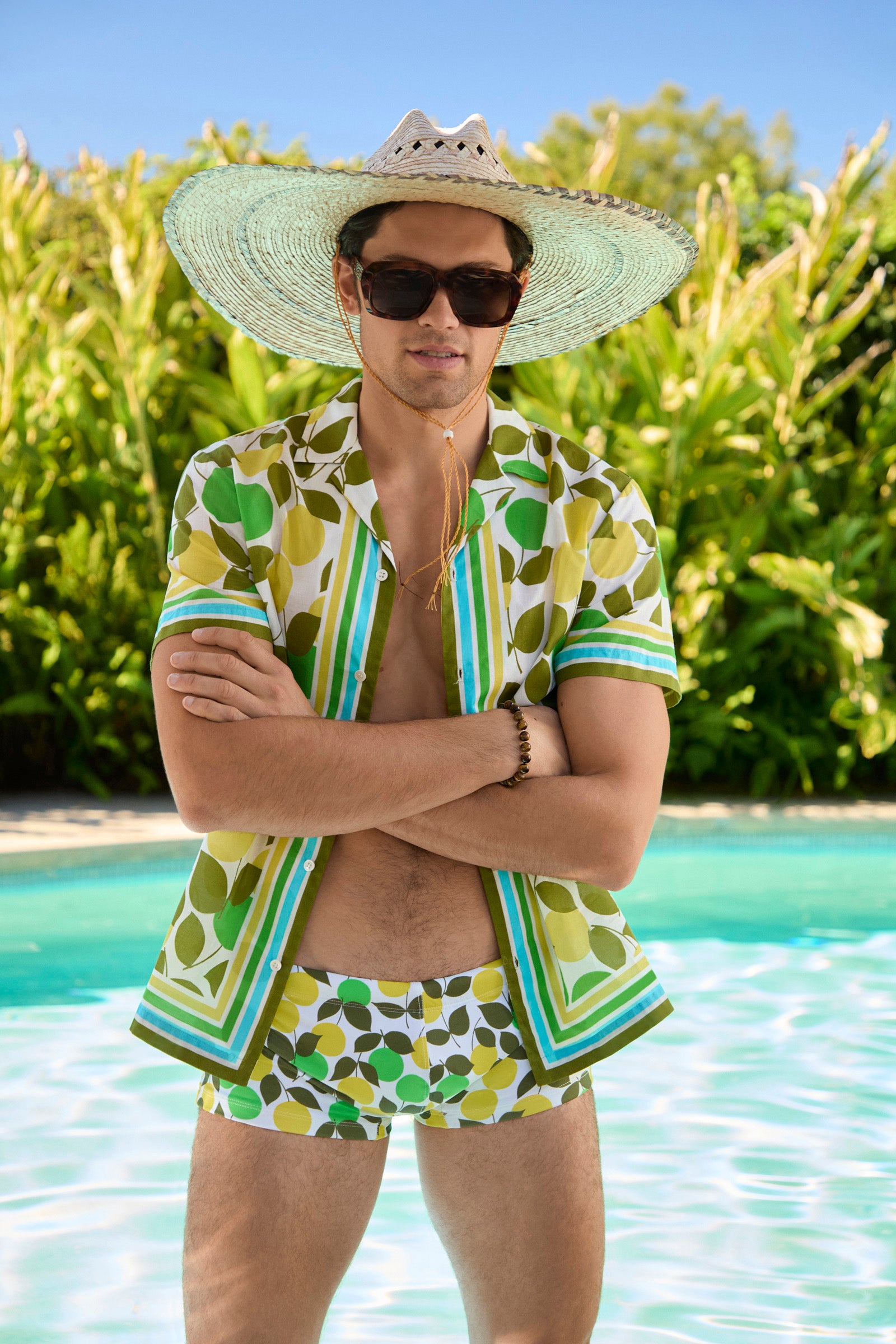 A man stands by a pool in the Sorrento Swim Trunk - Limone, paired with a matching patterned shirt, large straw hat, and dark sunglasses. He crosses his arms, surrounded by lush greenery in the background.