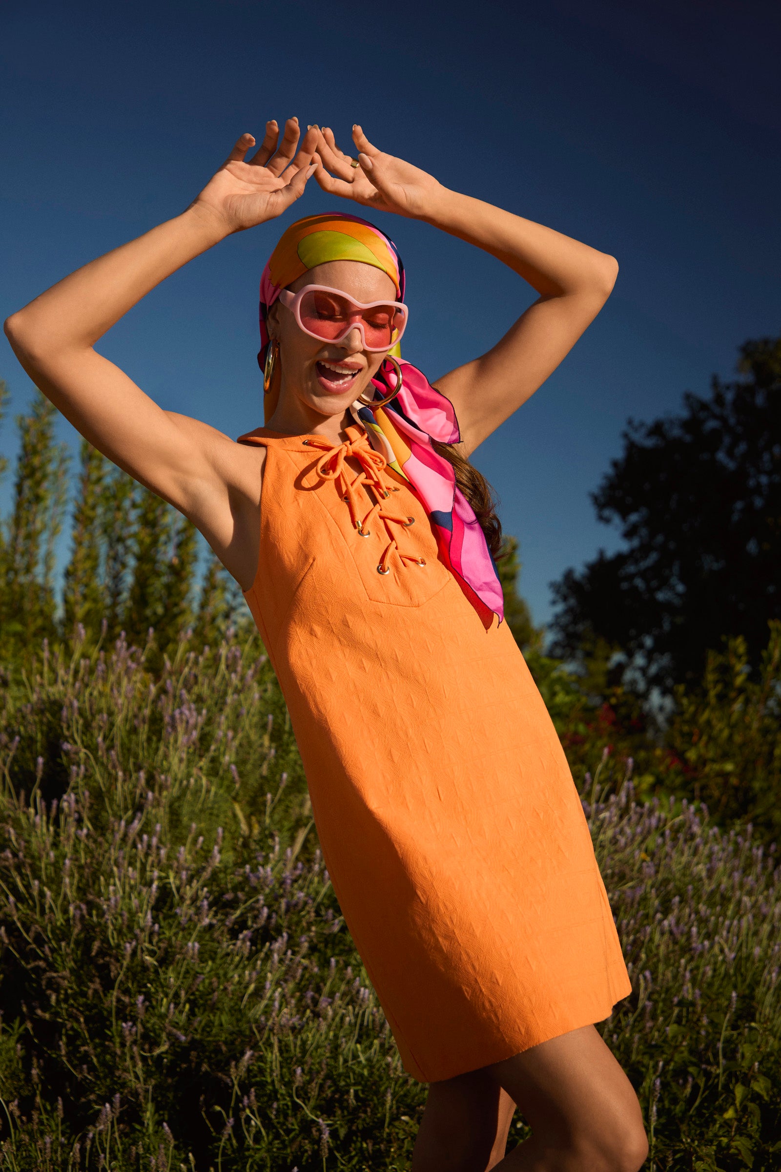 A joyful woman outdoors wears the Cappuccia Lace-Up Mini Dress in orange, paired with a colorful headscarf and pink sunglasses, raising her arms against a backdrop of blue sky and green plants.