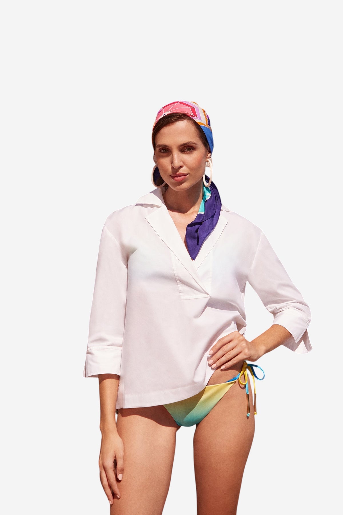 A woman stands outdoors on a sunny day in front of palm trees, wearing a white long-sleeve shirt, colorful bikini bottoms, large hoop earrings, and the Umbrella Scarf - Parapluie Silk as a vibrant headscarf.