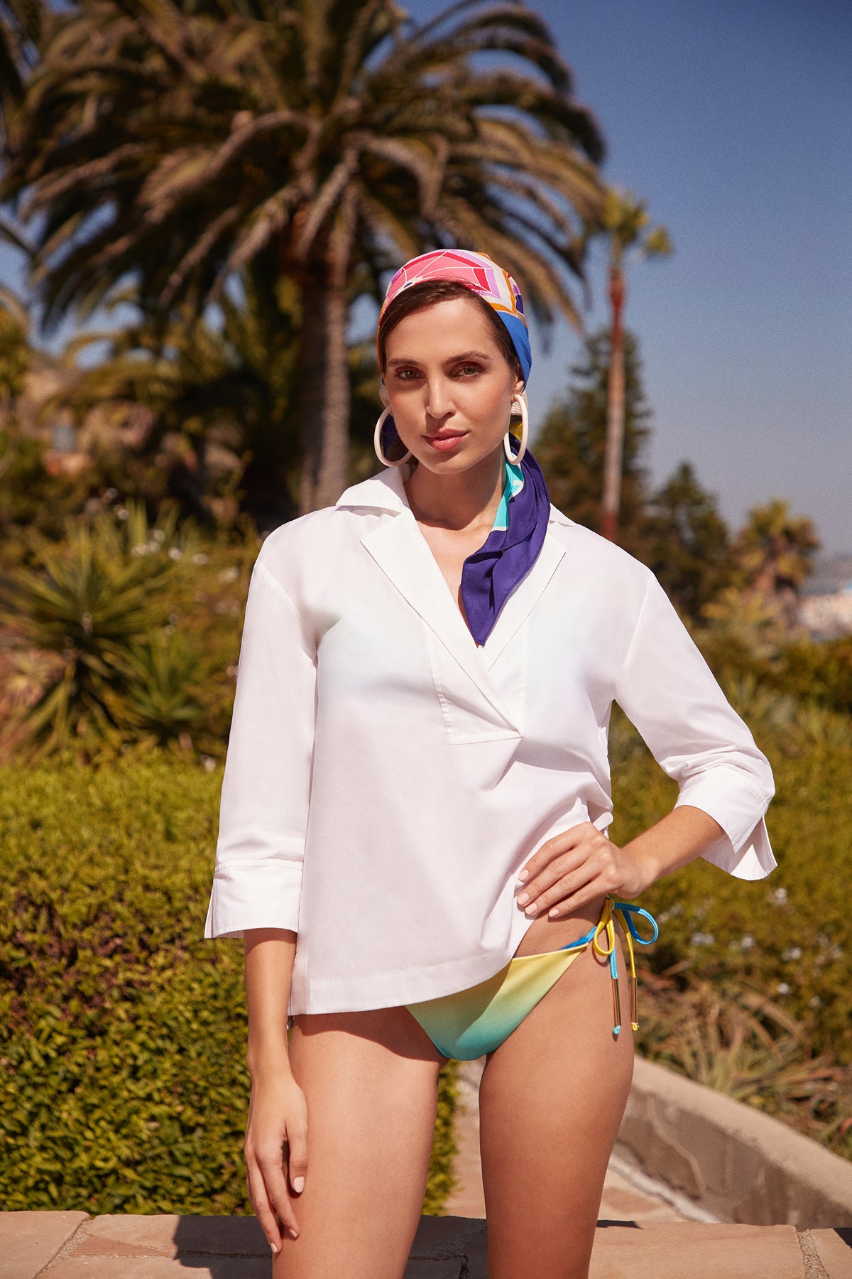 A woman stands outdoors on a sunny day in front of palm trees, wearing a white long-sleeve shirt, colorful bikini bottoms, large hoop earrings, and the Umbrella Scarf - Parapluie Silk as a vibrant headscarf.