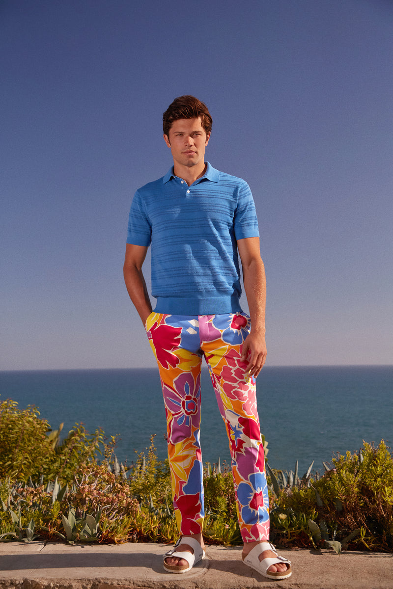 A man stands by the ocean in the Clyde Slim Trouser - Soie Paradis, featuring colorful floral prints, paired with a light blue polo shirt and white sandals. The clear blue sky, greenery, and water create a serene backdrop.