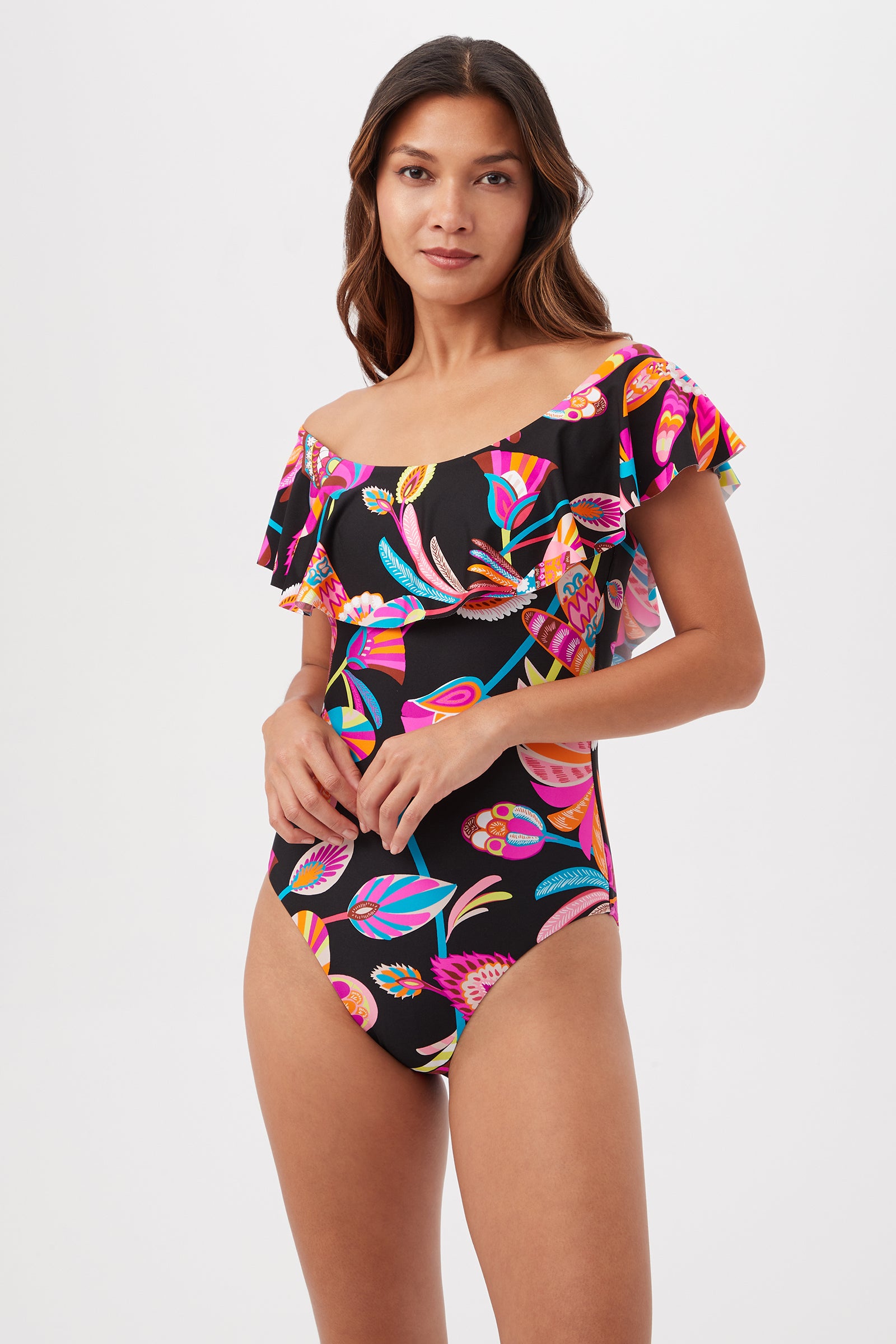 A woman with long brown hair models the Luminary Off-Shoulder One-Piece Swimsuit in Multi, featuring a ruffle detail and tropical floral pattern on black. She stands against a plain white backdrop, looking at the camera.