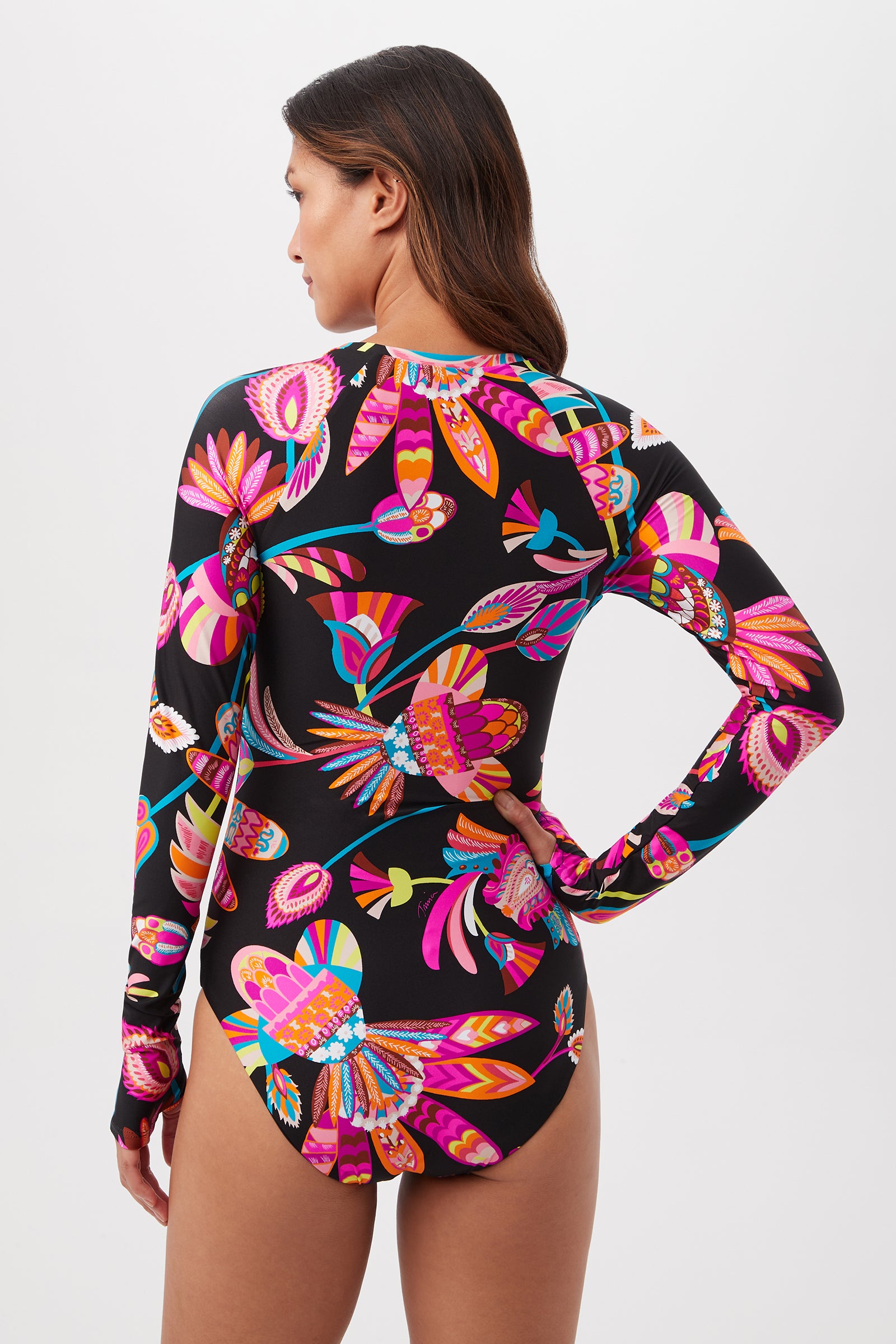 A woman with long brown hair models the Luminary Long-Sleeve Paddle Suit, showcasing its vibrant floral and abstract patterns as she faces away from the camera against a plain white background.