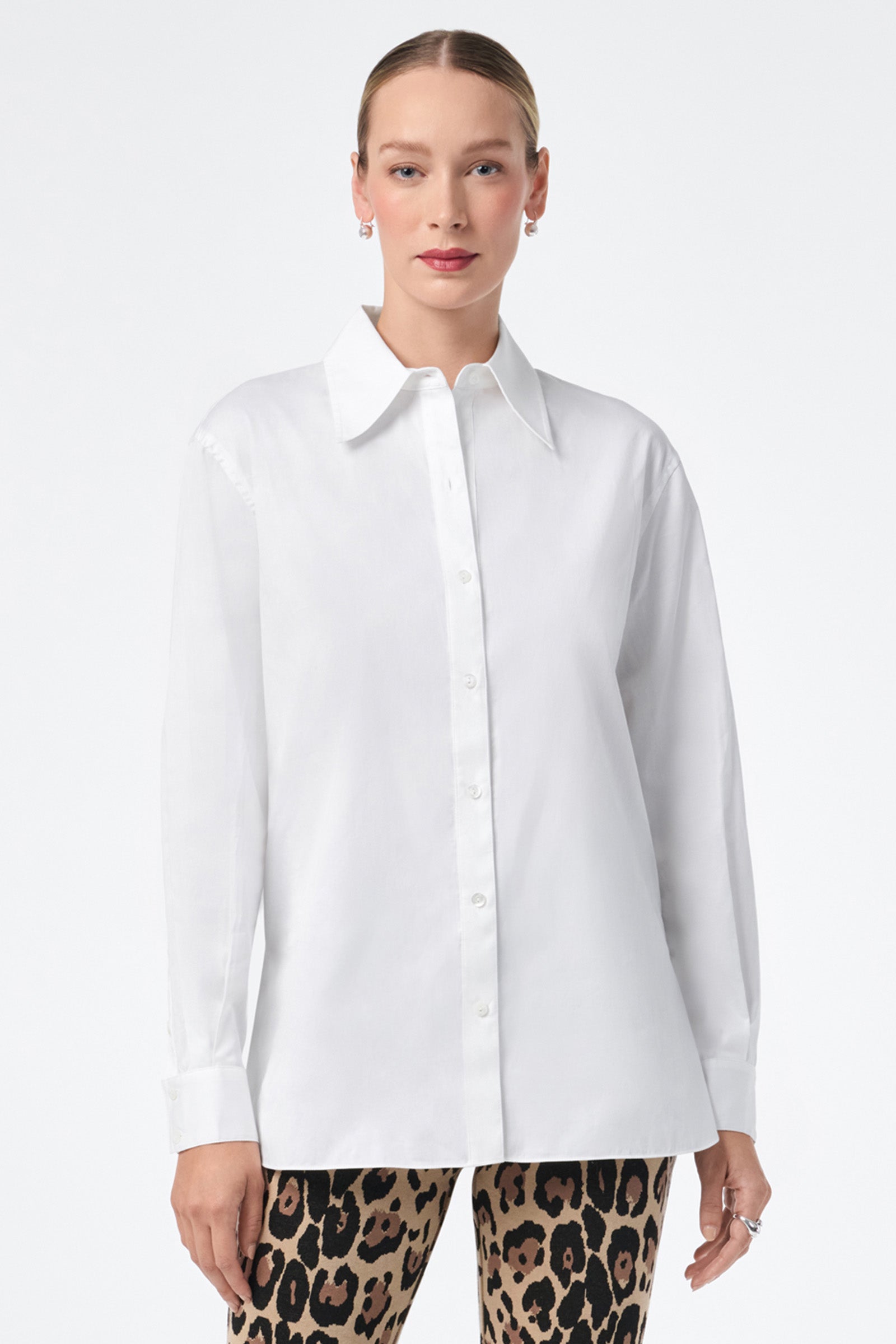 A woman poses against a plain background wearing the Litsa Button-Up Cotton Shirt, featuring a crisp white design with a pointed collar and long sleeves, paired with high-waisted leopard print pants.