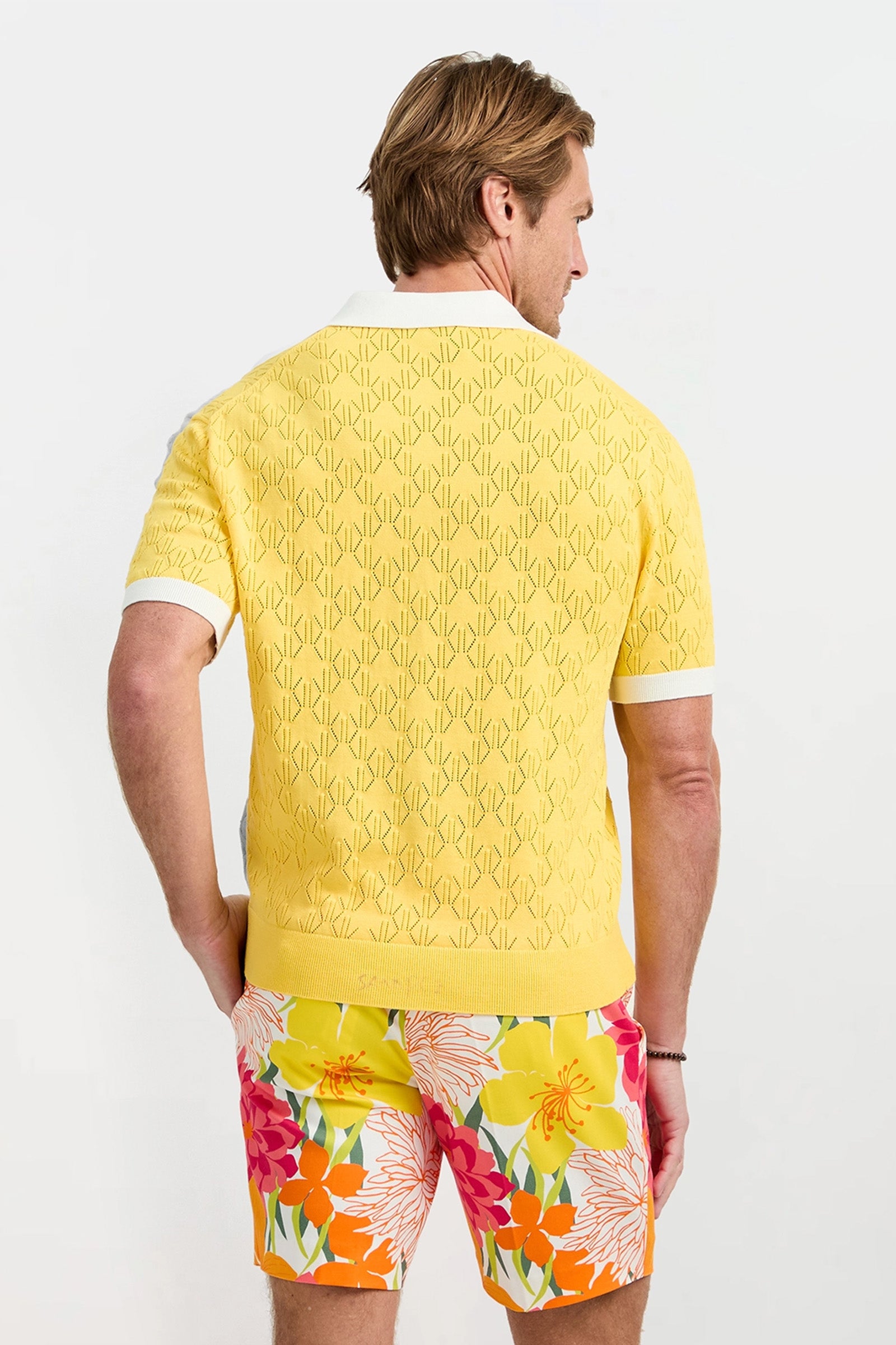 A man with light brown hair faces away, wearing the Luca Short-Sleeve Polo in a yellow pattern and bright floral shorts. The background is plain white.