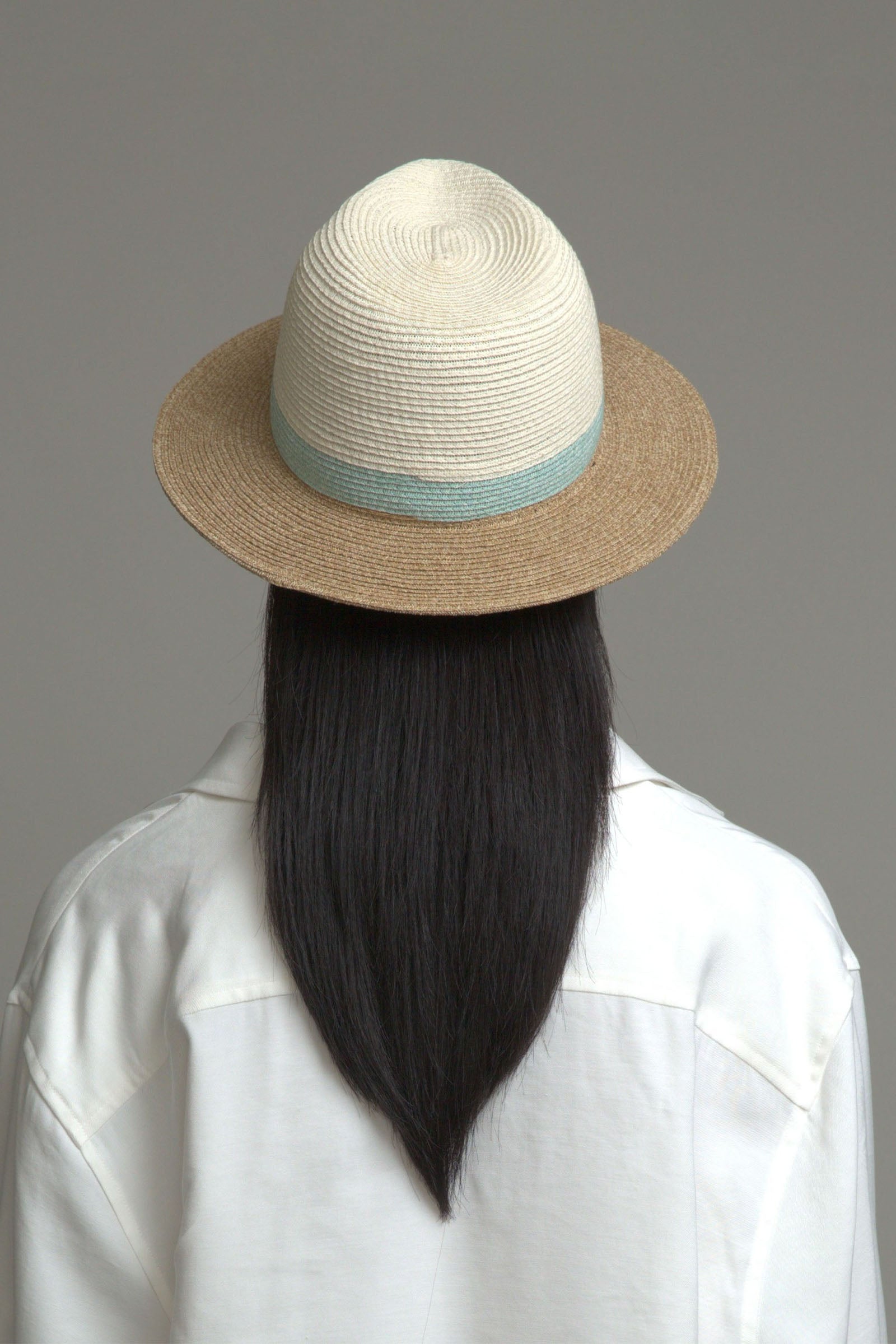 A person with long, straight black hair wears the Eugenia Kim Lyle Hat—a cream and brown packable fedora with a light blue band—paired with a white collared shirt, shown from behind against a plain gray background for effortless beach-day style.