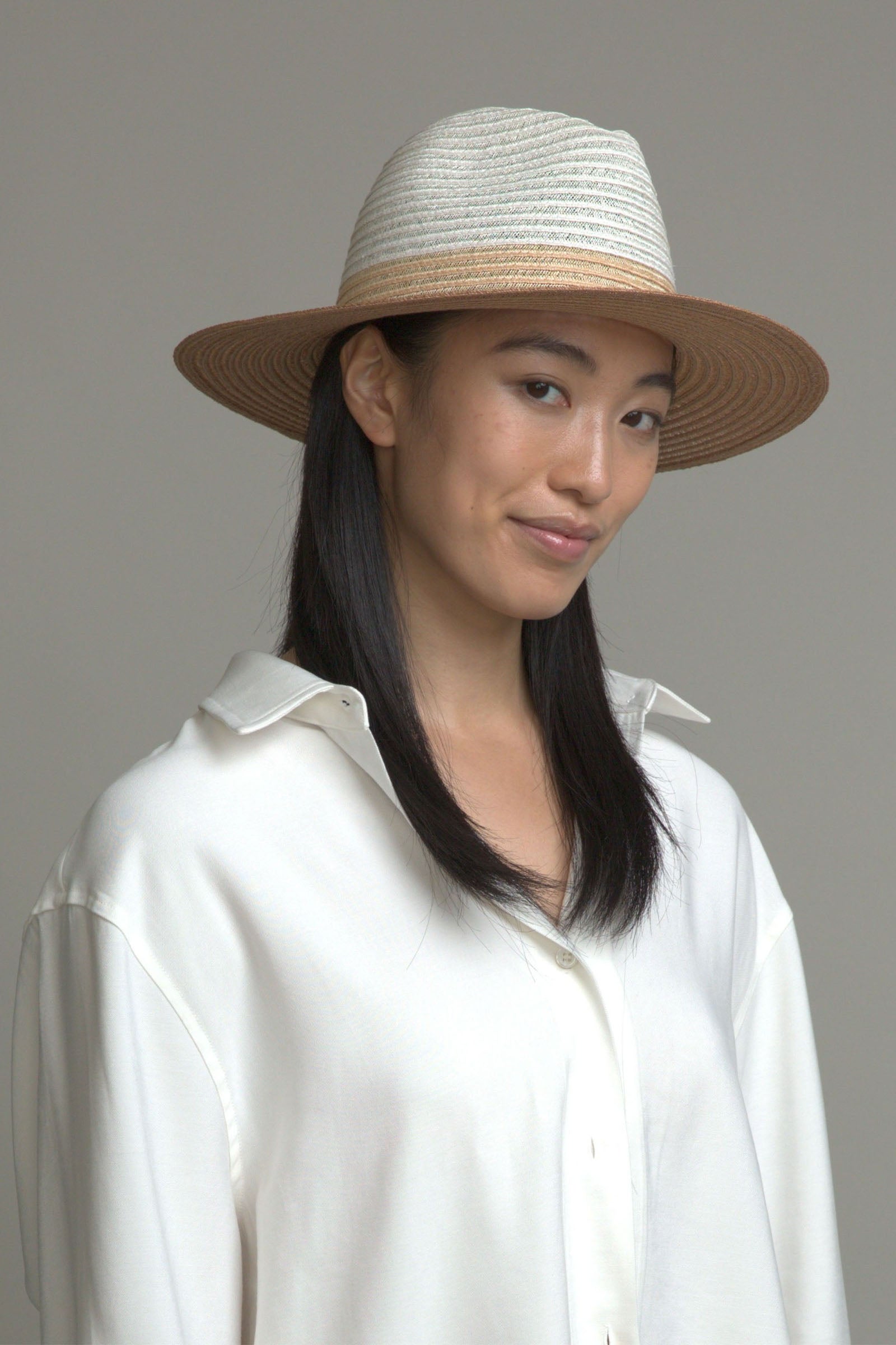 Wearing the Eugenia Kim Lyle Hat and a loose white shirt, a woman with straight black hair stands against a gray background, smiling softly and capturing effortless beach-day style.
