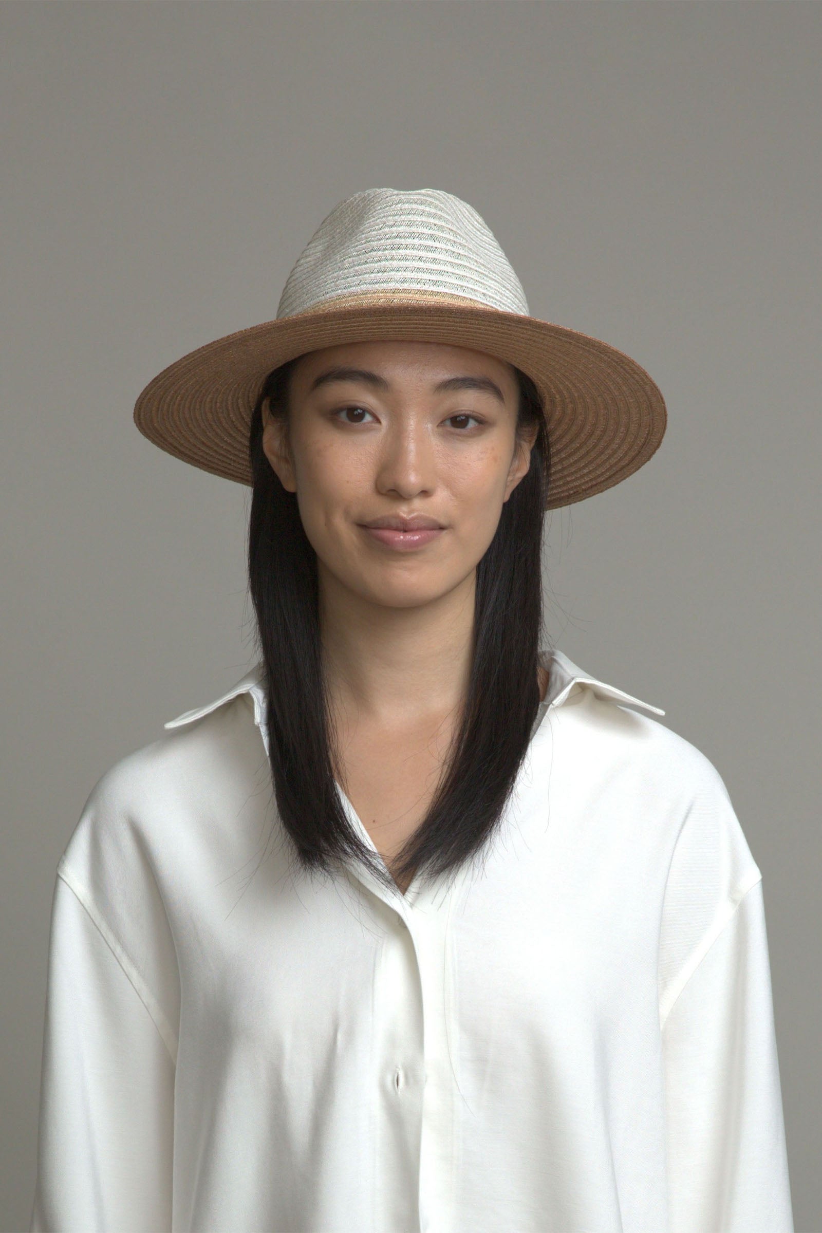 A woman with straight black hair smiles softly while wearing the Eugenia Kim Lyle Hat and a loose white shirt, standing against a plain gray background.