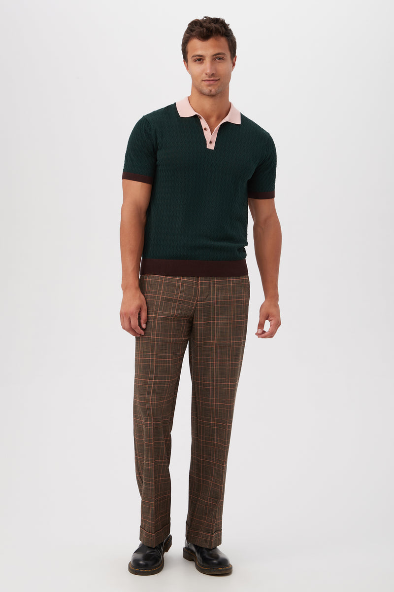 A man stands against a white background, facing forward with a relaxed expression, wearing Genet Wide-Leg Trousers - Chaka Check Plaid, a dark green short-sleeve polo with a pink collar, and black shoes.