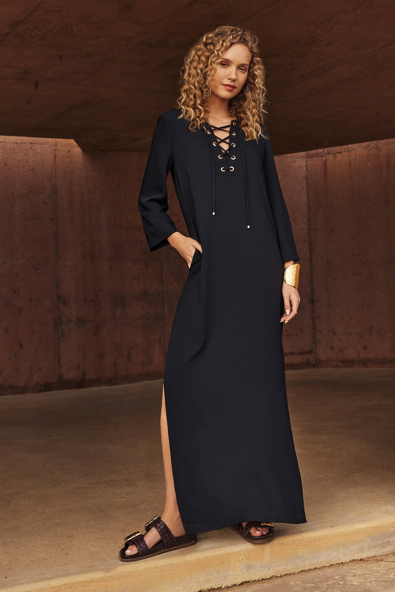 A woman with curly blonde hair wears the Catamaran 3/4-Sleeve Maxi Dress in black, paired with black sandals and a gold cuff bracelet, standing against a brown concrete background.
