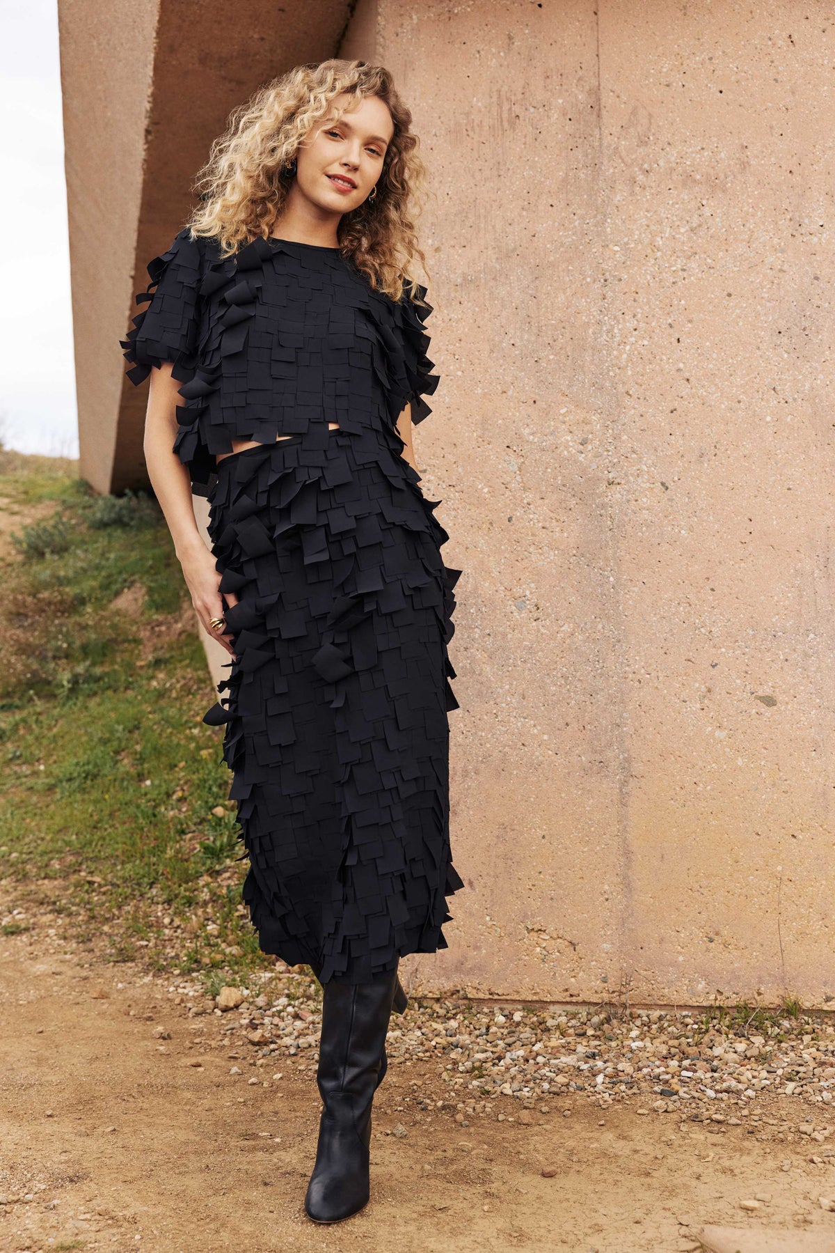 A woman with curly blonde hair stands outdoors in front of a concrete structure, wearing the Serilda Short-Sleeve Top - Flutter Chiffon with black knee-high boots.