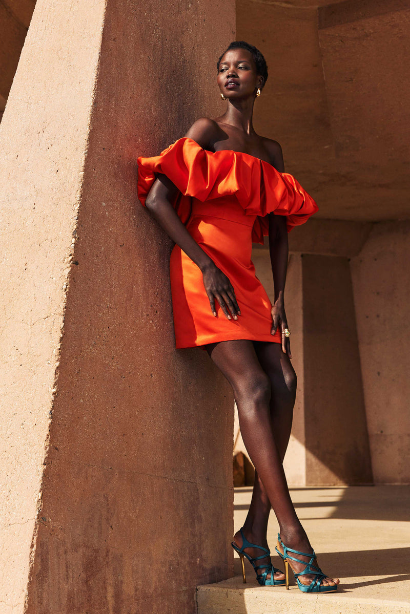 A woman in the Nia Satin Off The Shoulder Short Cocktail Dress stands against a tan, textured structure, looking confidently into the distance.