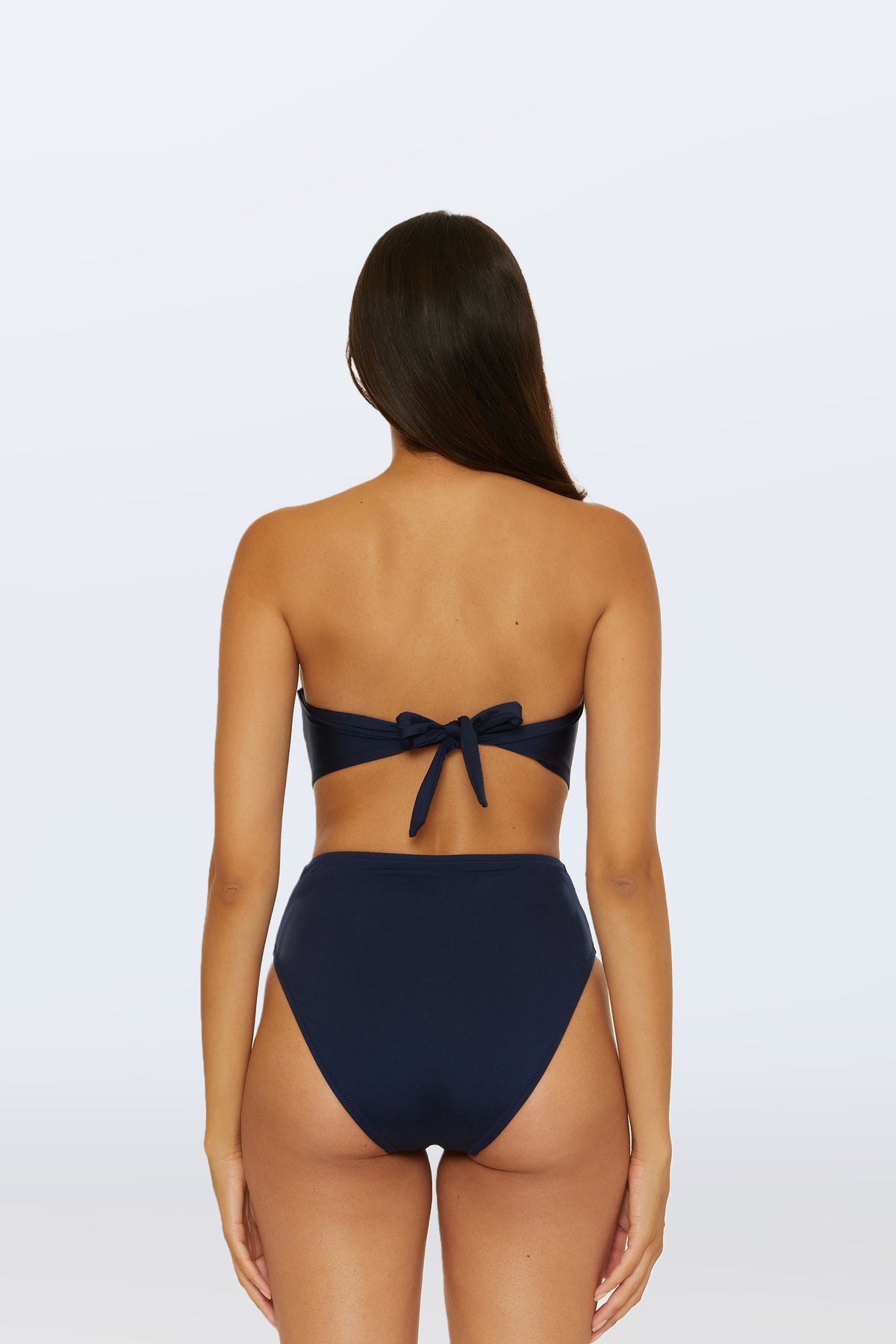 A woman with long brown hair is shown from behind, wearing the Monaco Pleated Tab Bandeau Swim Top in navy blue with a high-waisted bottom. The bandeau top features a bow tie at the back against a plain light gray background.