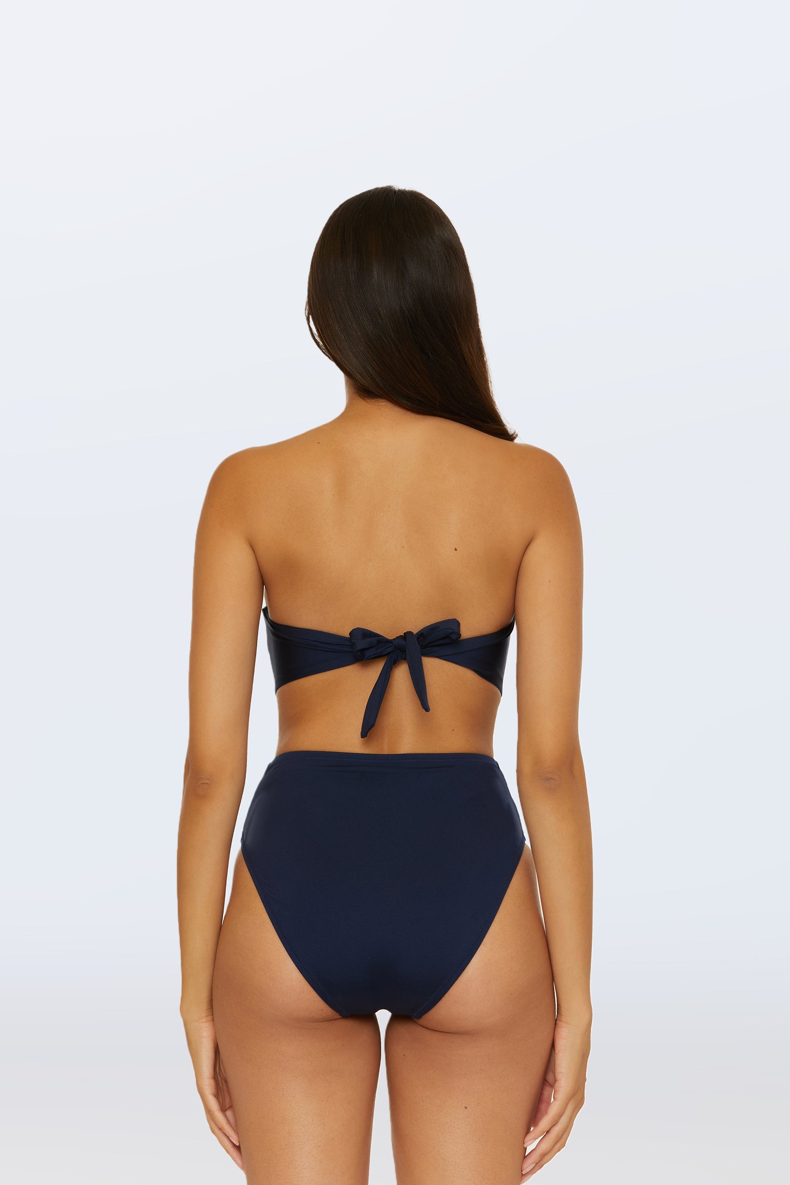 A woman with long brown hair is shown from behind, wearing the Monaco Pleated Tab Bandeau Swim Top in navy blue with a high-waisted bottom. The bandeau top features a bow tie at the back against a plain light gray background.