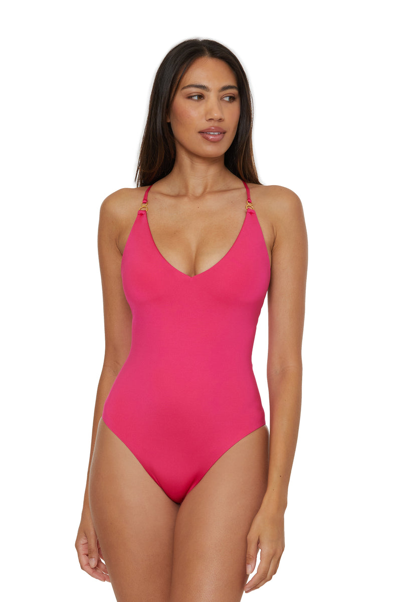 A woman in the Monaco Twist Back One-Piece Swimsuit in Cannes Coral, featuring thin straps and gold accents, stands against a plain white background, looking slightly to the side.