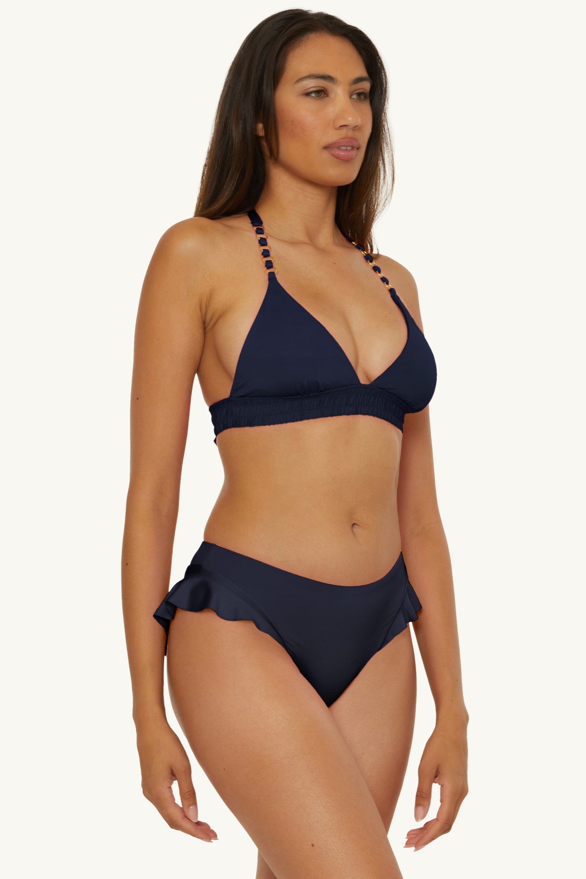 A woman wearing the Ring Halter Swim Top - Monaco, a navy blue bikini with beaded straps and ruffle detail bottoms, stands against a plain light background.