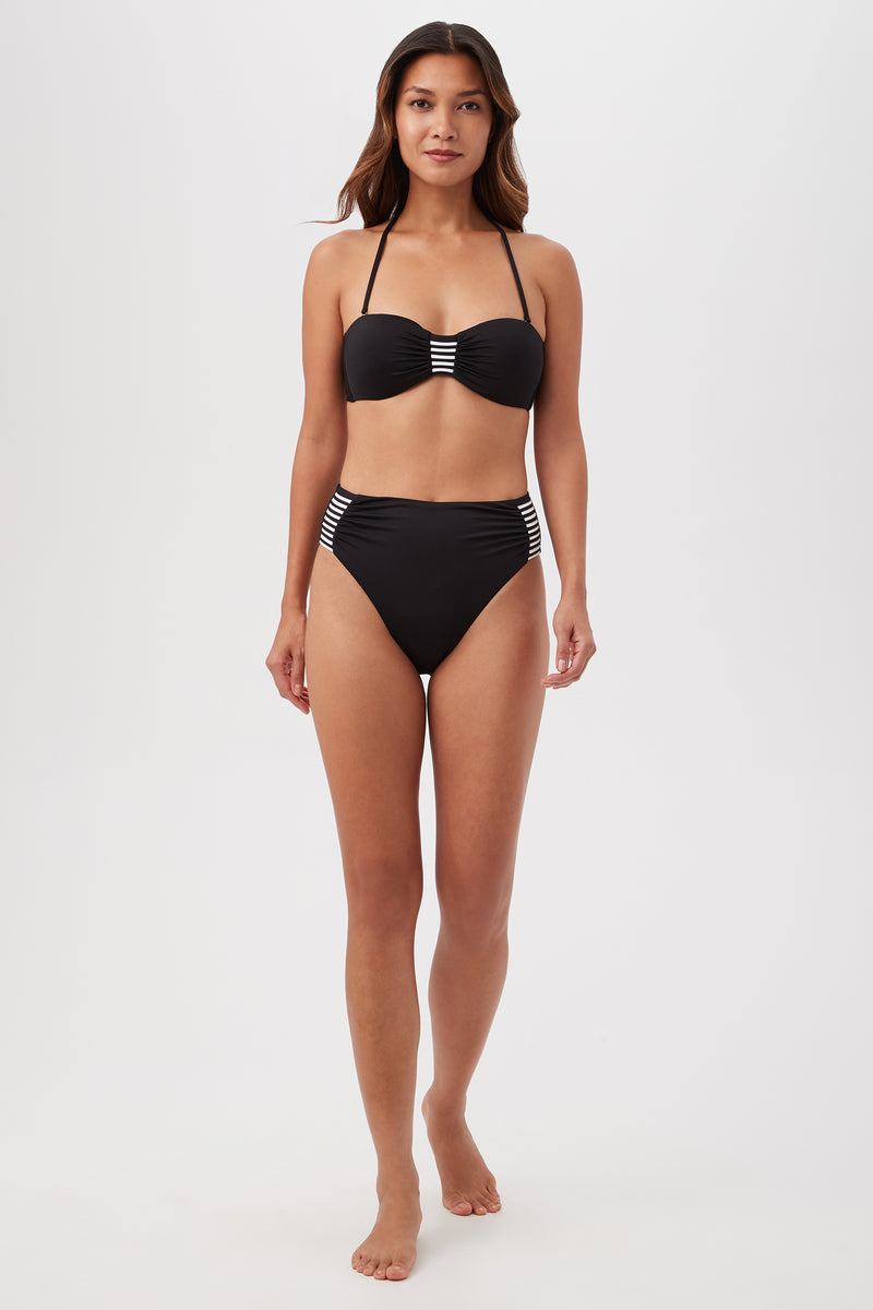 A woman with long, wavy brown hair models the Monaco Pleated High-Waist Bikini Bottom, featuring black fabric with white stripes, against a plain white background. She faces forward with a neutral expression and relaxed arms.