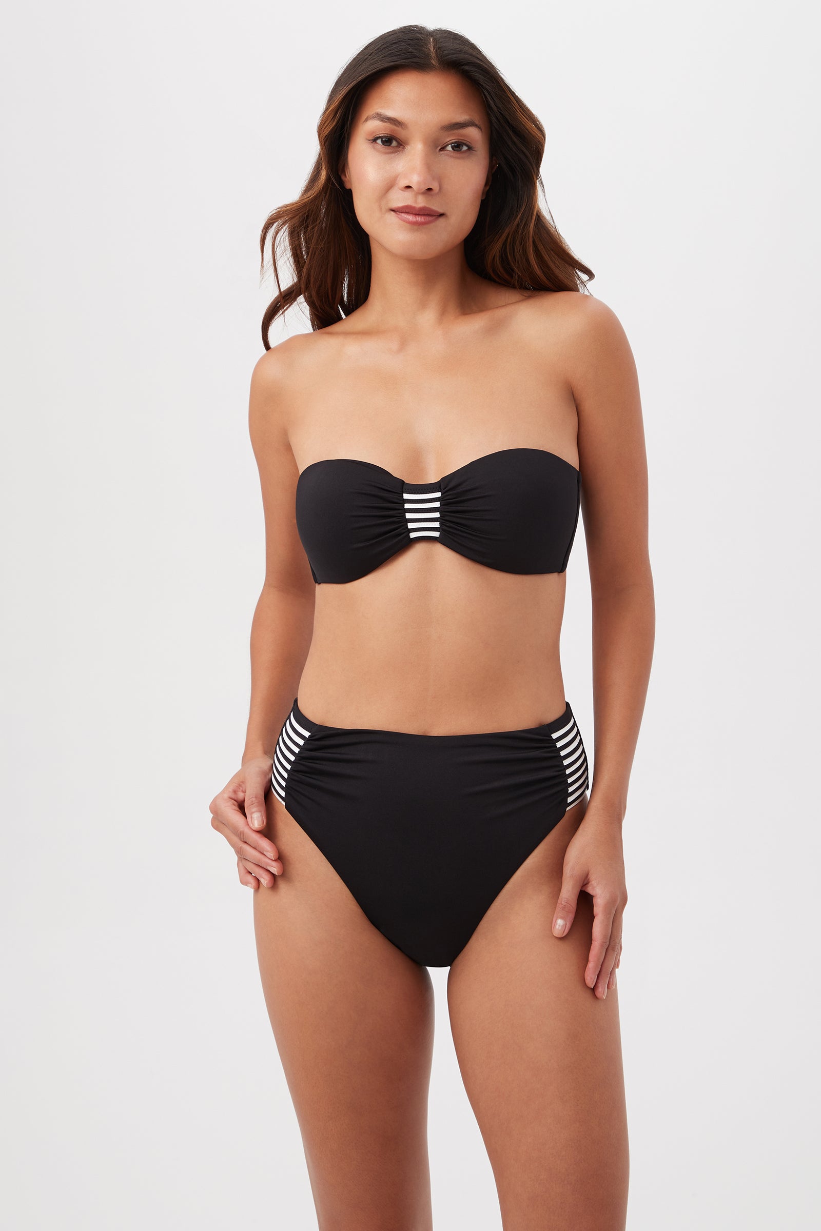 A woman wears the Monaco Pleated Tab Bandeau Swim Top in black/white, featuring striped accents, as she poses against a plain light background.