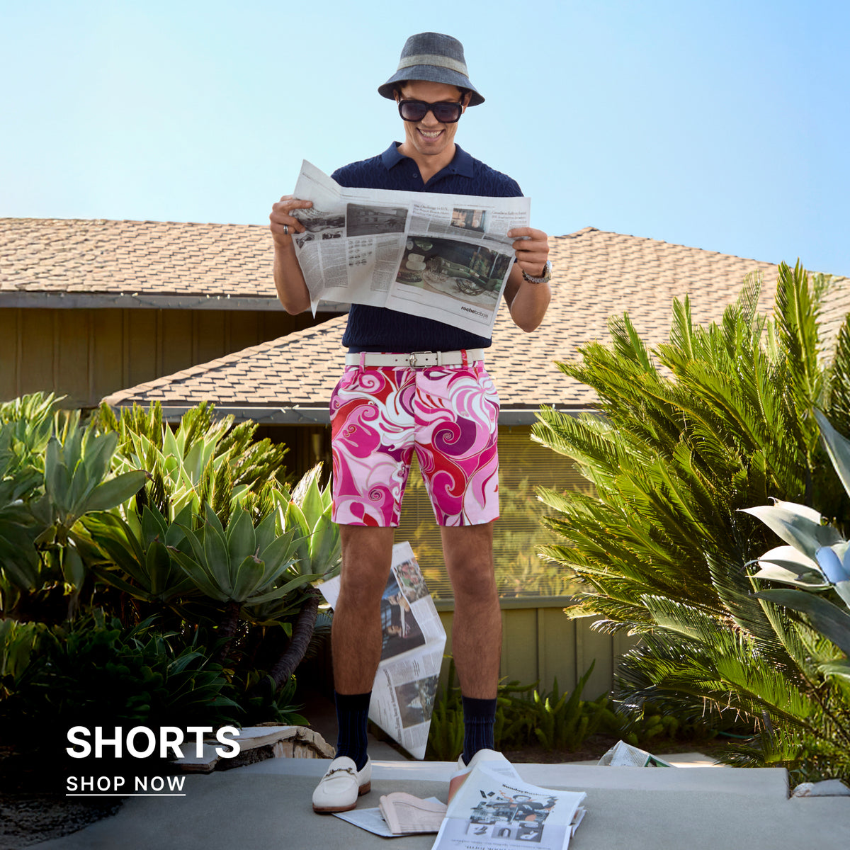 Man wearing colorful shorts and a hat, holding a newspaper outdoors.