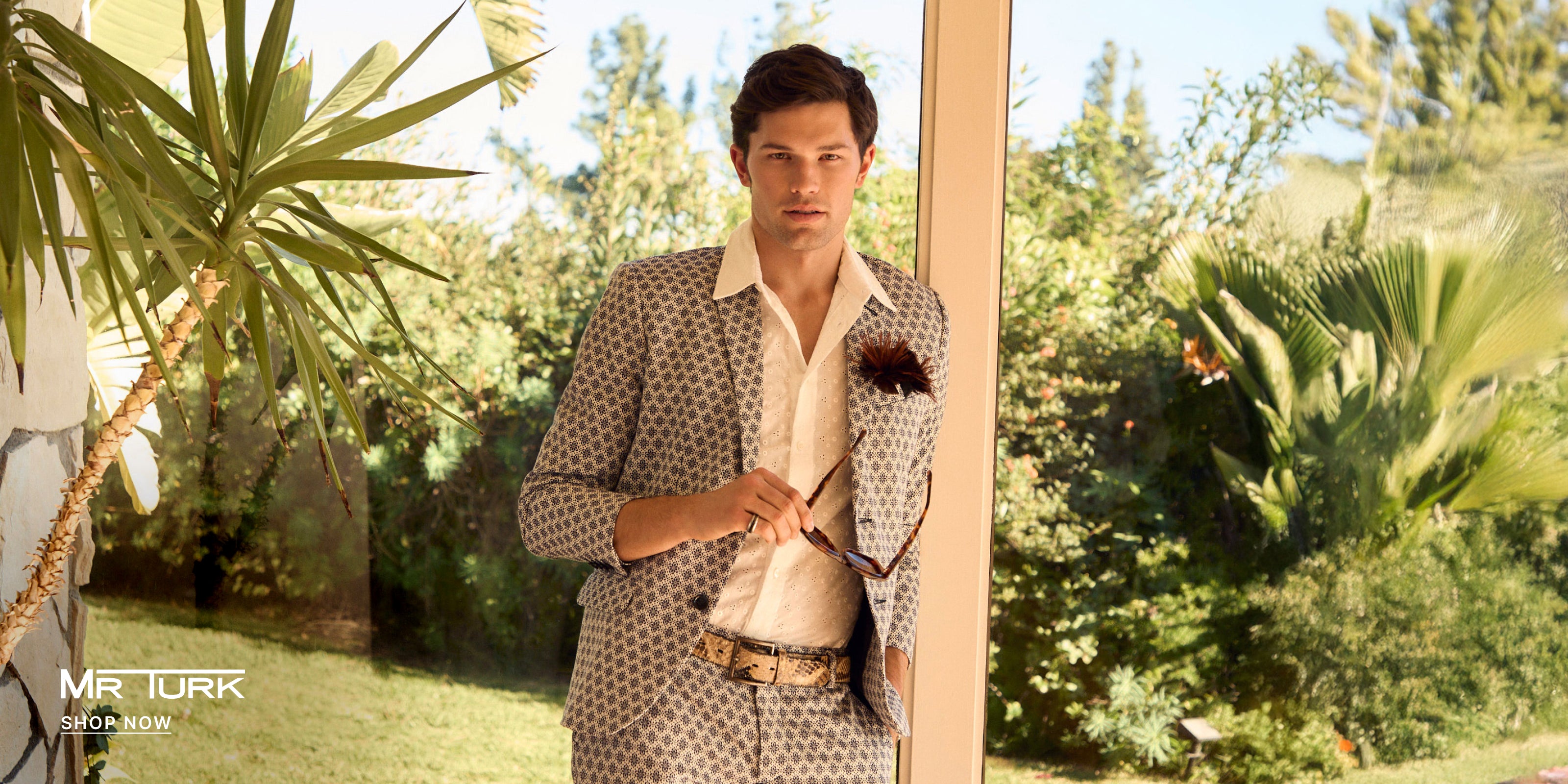 Man in a patterned suit standing outdoors with greenery in the background