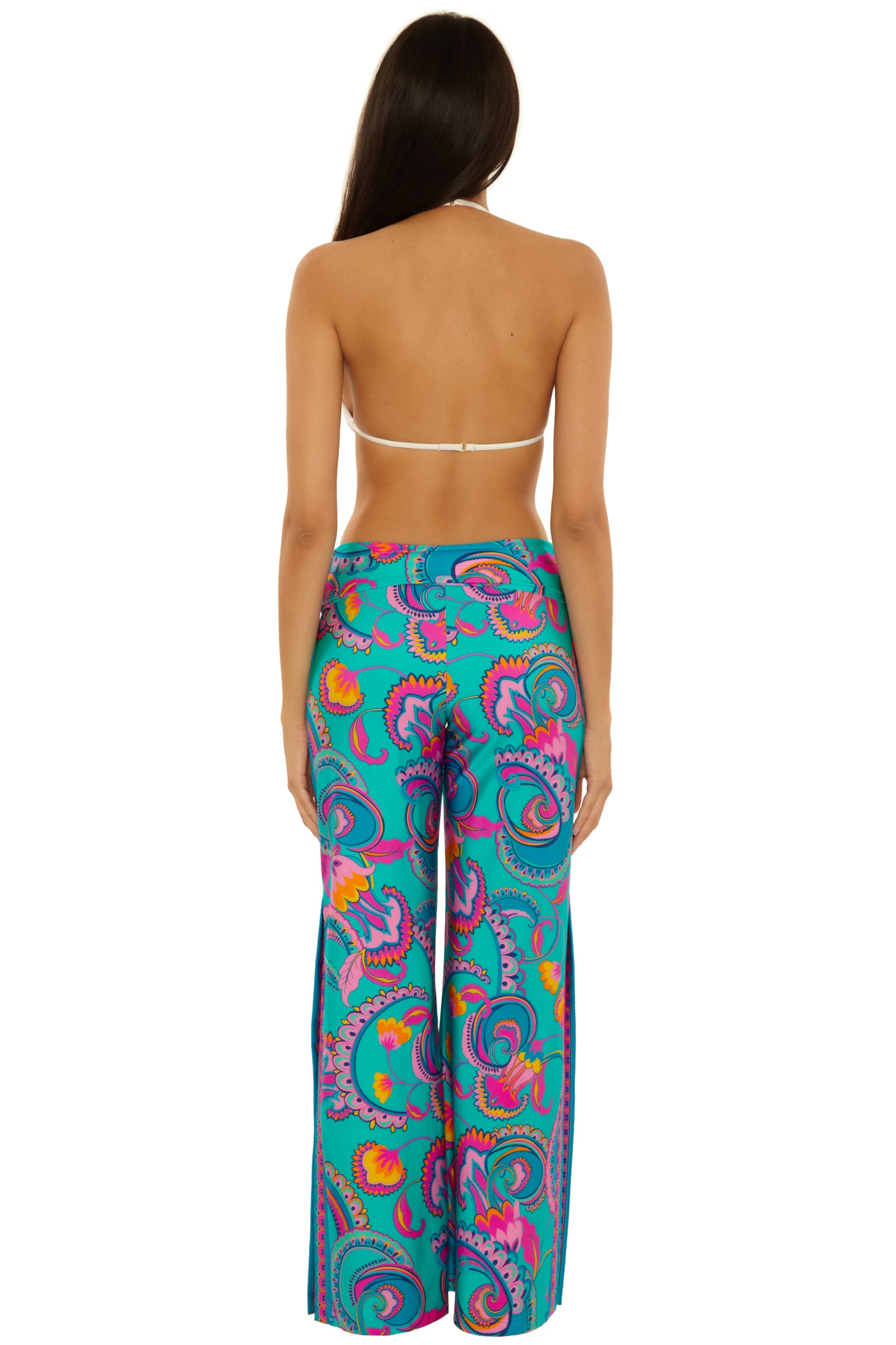 A woman with long brown hair faces away, wearing a white halter bikini top and the Mykonos Border Swim Pant—high-waisted, wide-leg pants featuring a vibrant paisley pattern in blue, pink, orange, and purple.