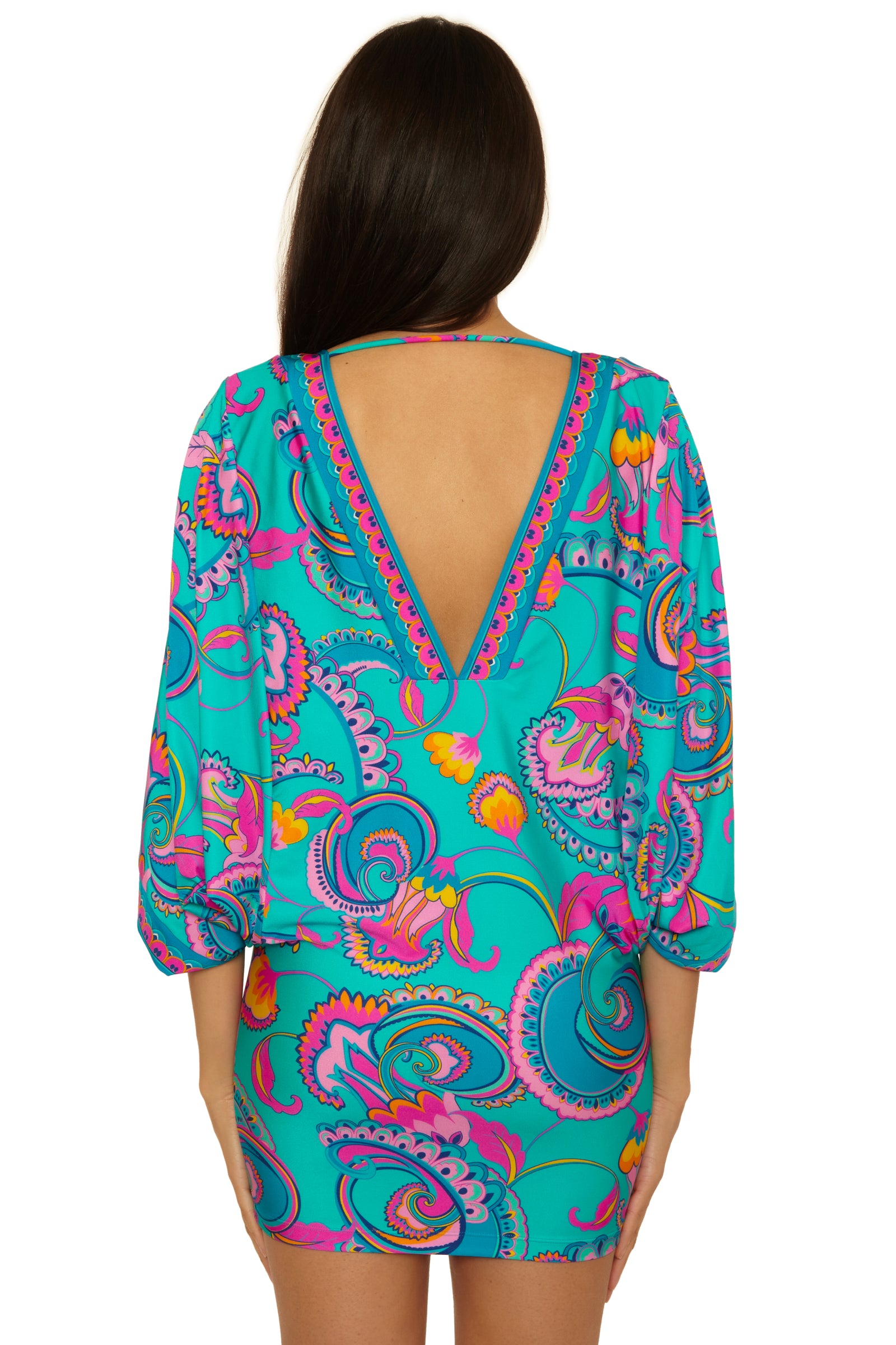 A woman with long brown hair stands facing away, wearing the Mykonos Casablanca Tunic—a vibrant blue dress featuring a colorful paisley pattern and a deep V-cut open back.