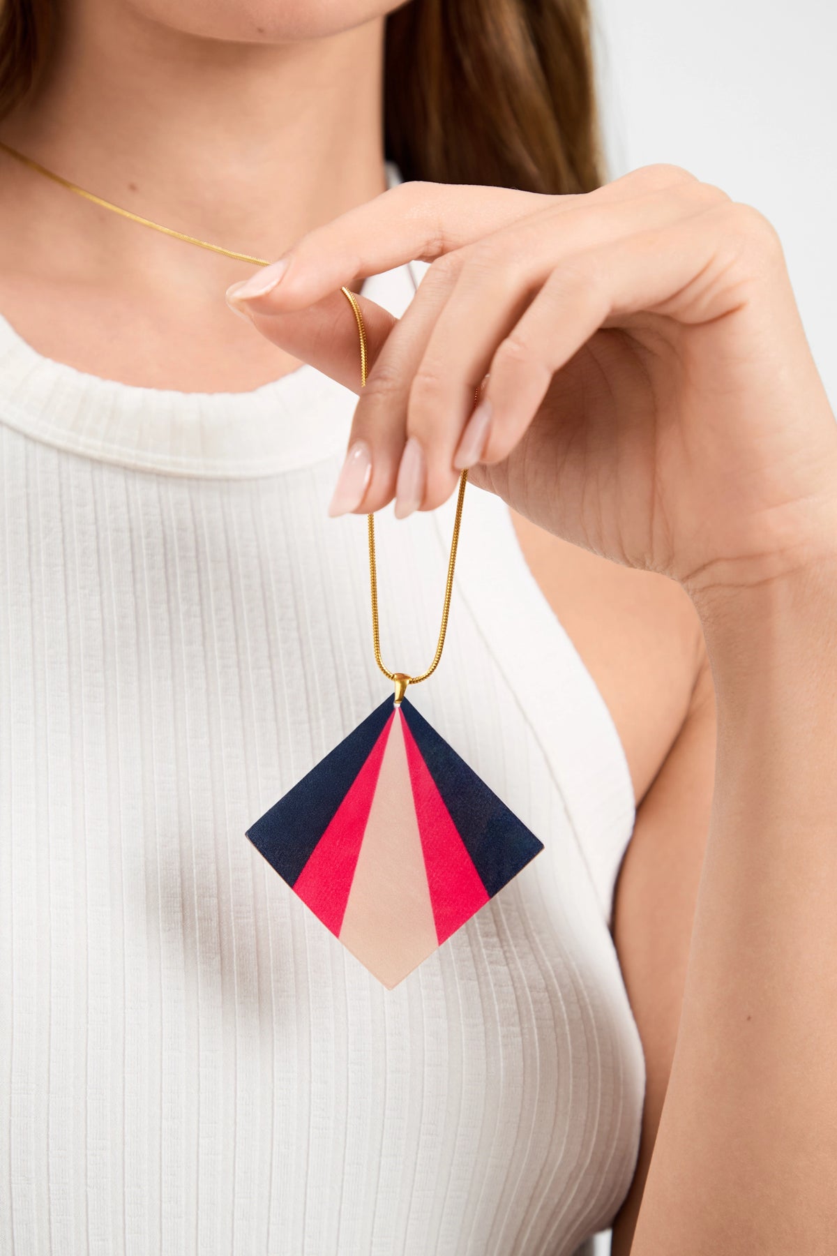 Wearing a white sleeveless top, a person holds the Kite Necklace, featuring geometric navy blue, red, and beige color blocks on a pendant suspended from a thin gold chain.