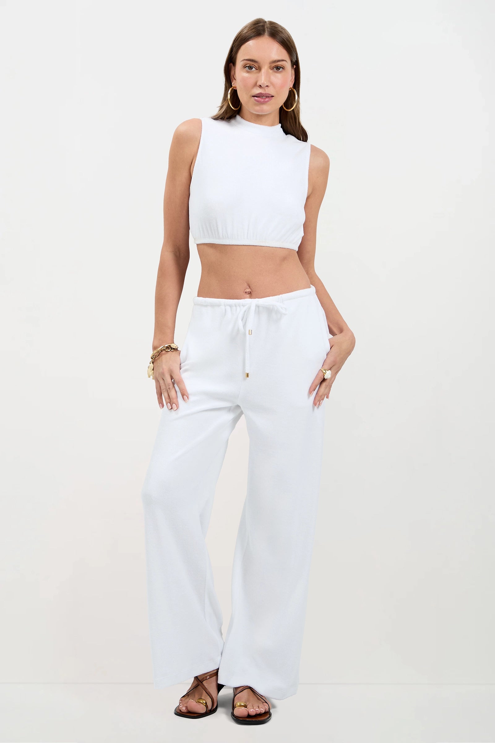 A woman poses against a plain background in the Linsk Wide-Leg Pant - Poolside Terry, paired with a sleeveless white crop top. She completes the look with gold jewelry and flat sandals.